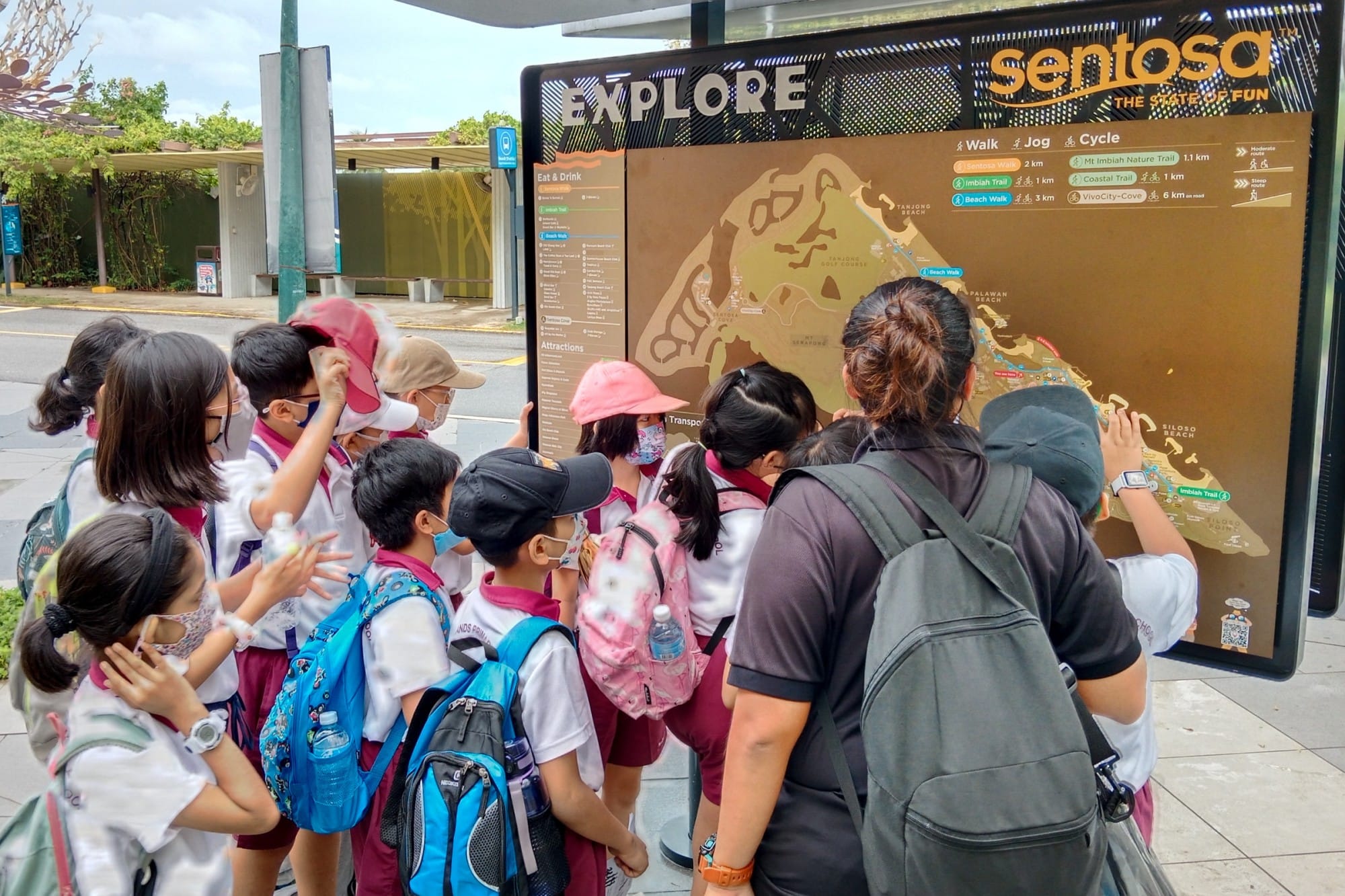 A group of school children with backpacks gather around a Sentosa island map board labeled Explore.