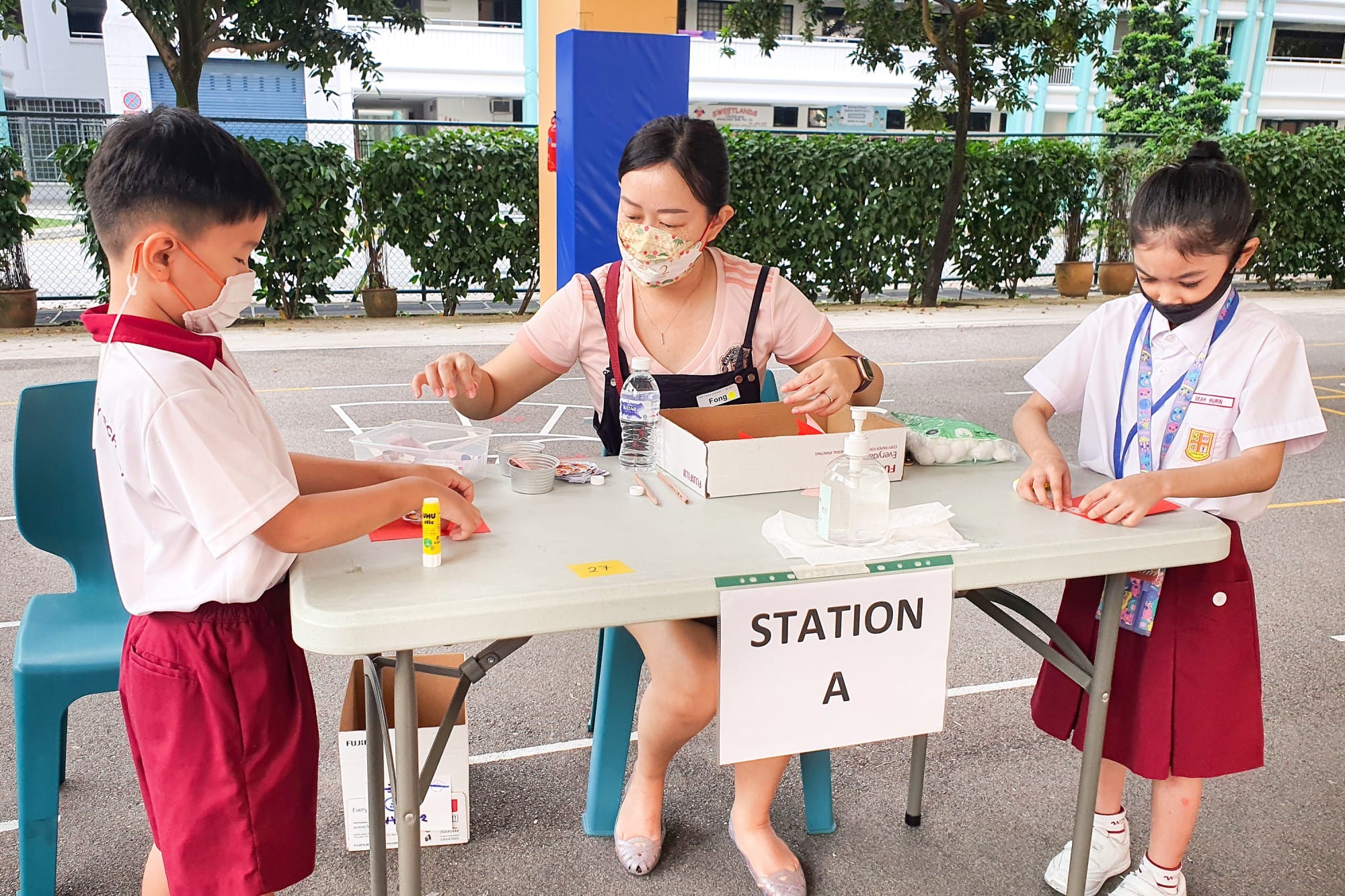 Two masked children in school uniforms do a craft activity at a table labeled Station A, supervised by a masked adult.