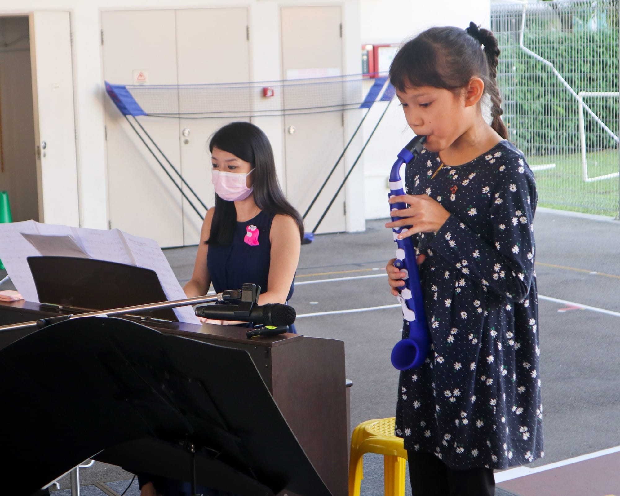 A young girl plays a blue recorder while a masked woman accompanies her at a keyboard with sheet music.