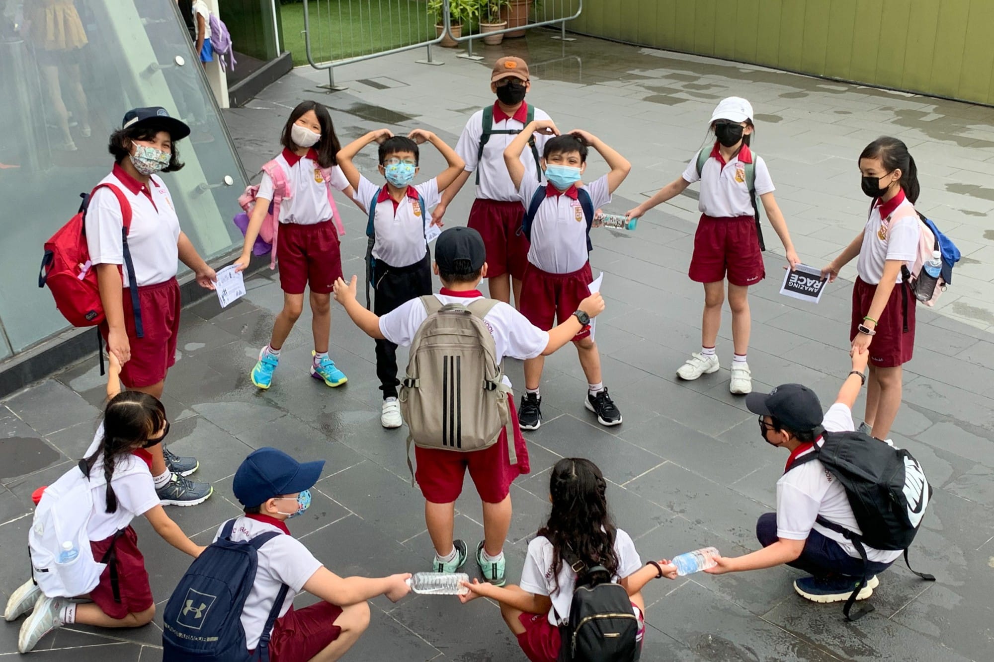 A group of masked children in white and red school uniforms with backpacks play a game outdoors on a paved area.