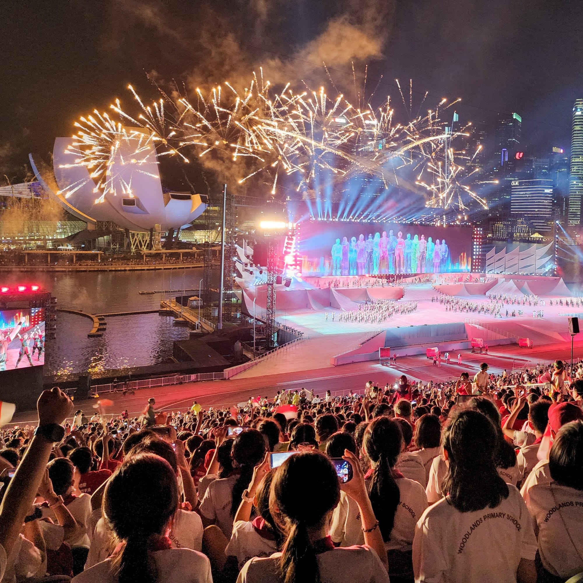 Large crowd watches a night ceremony with golden fireworks, colorful stage lights, and a city skyline near a waterfront.