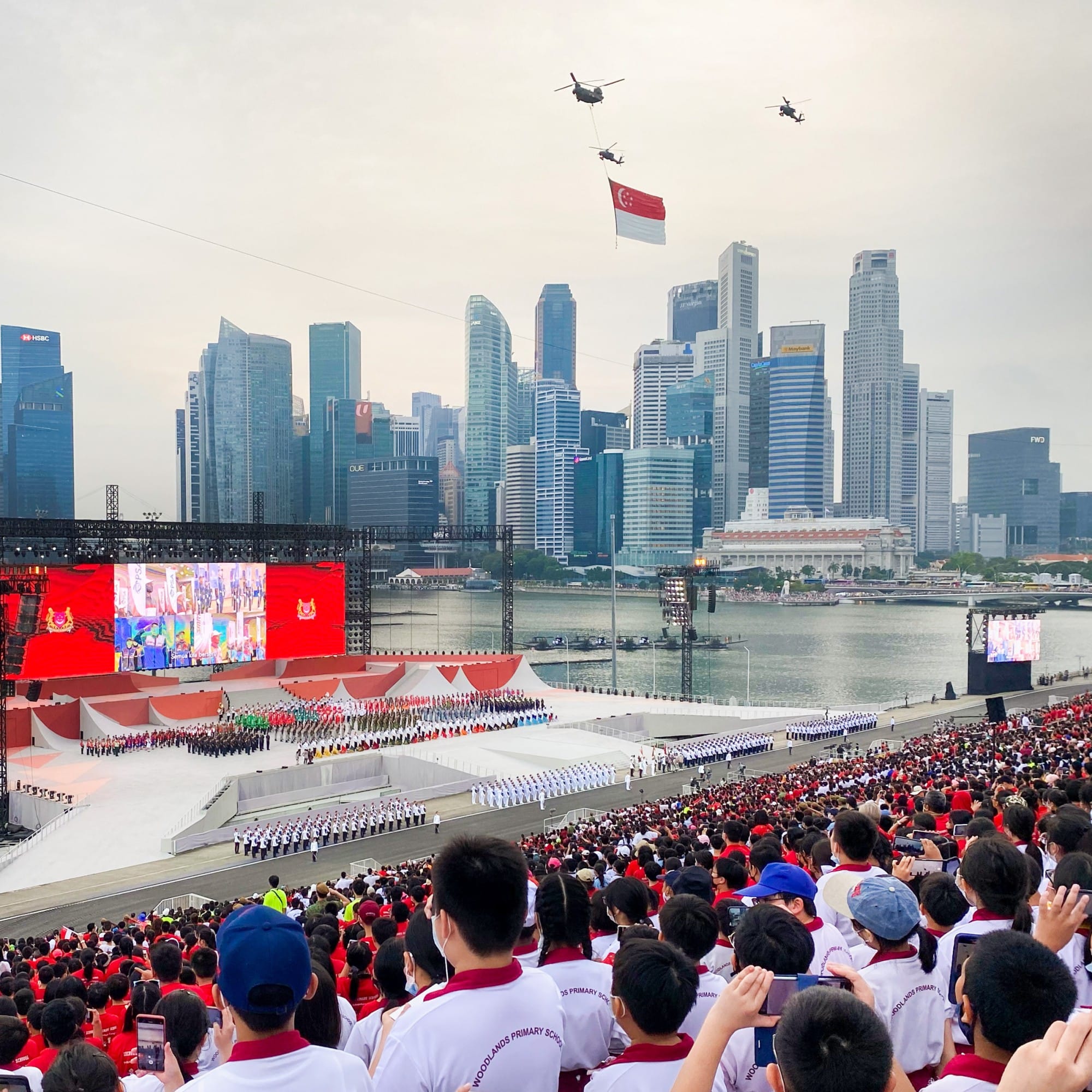Helicopters carry the Singapore flag over a large outdoor parade with crowds, marching formations, and a city skyline backdrop.