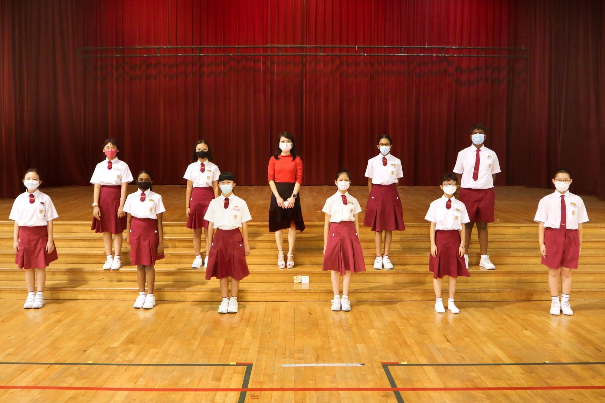 A teacher in red top stands with 11 masked students in white and dark red uniforms on a school hall stage.