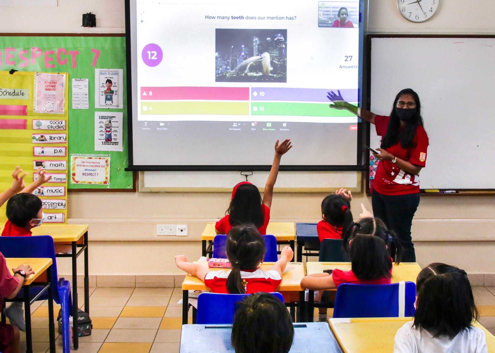 A masked teacher points at a quiz slideshow while young students in red uniforms raise their hands in a colorful classroom.