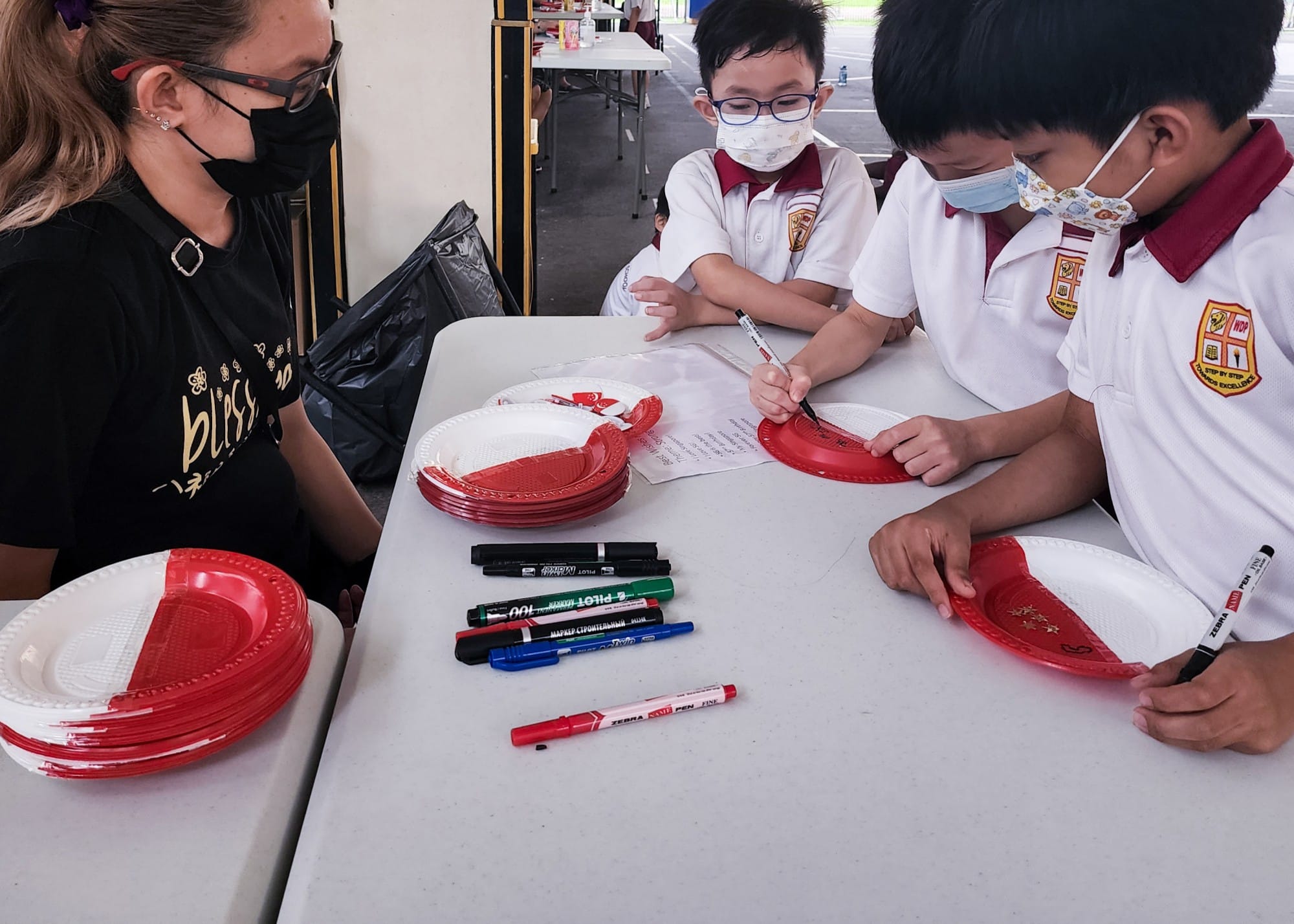Two masked schoolboys in uniforms draw on red and white paper plates with markers at a table supervised by a masked adult.
