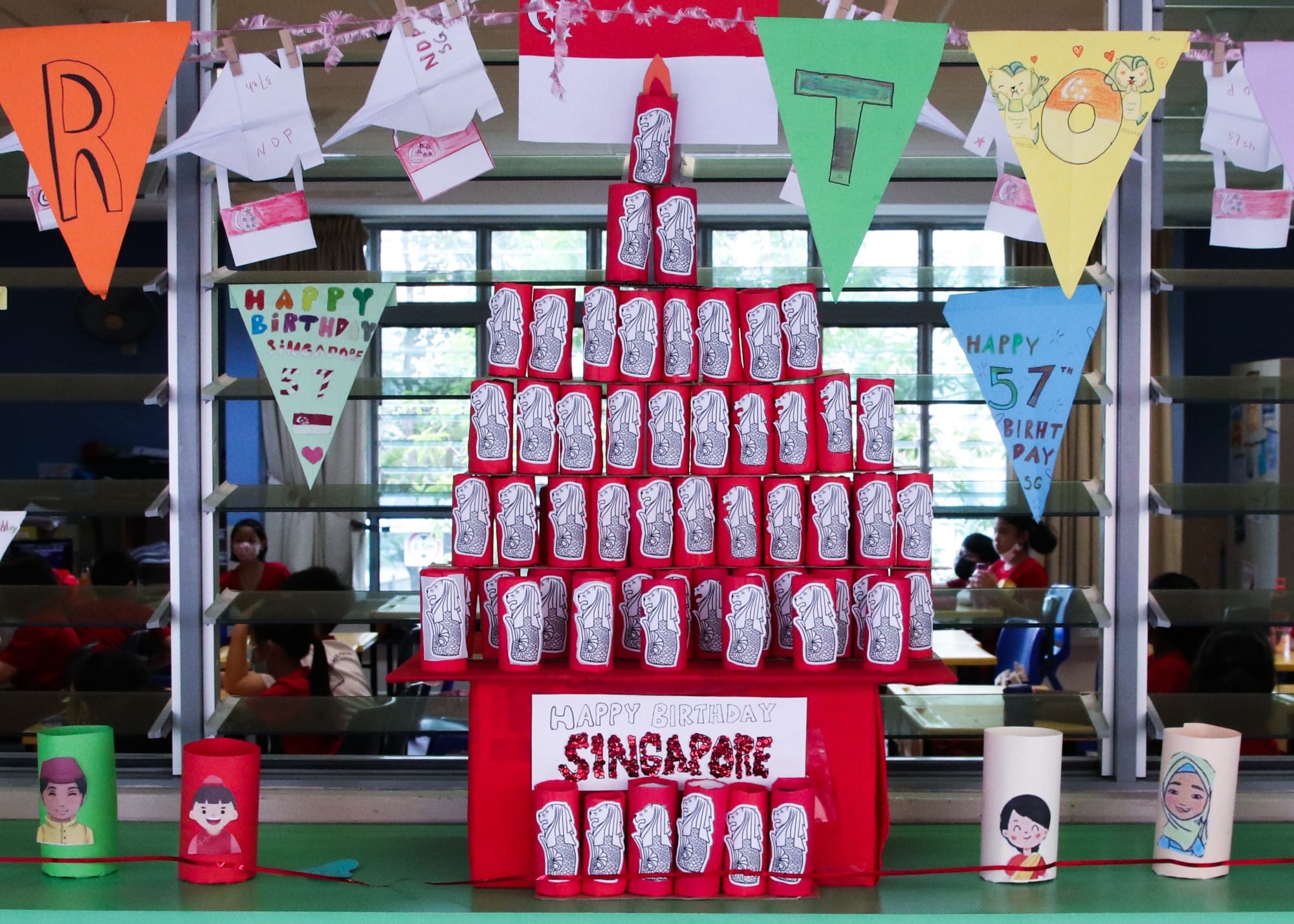 A pyramid of red Merlion-decorated cups with a Happy Birthday Singapore sign and colorful bunting celebrating Singapore's 57th birthday.