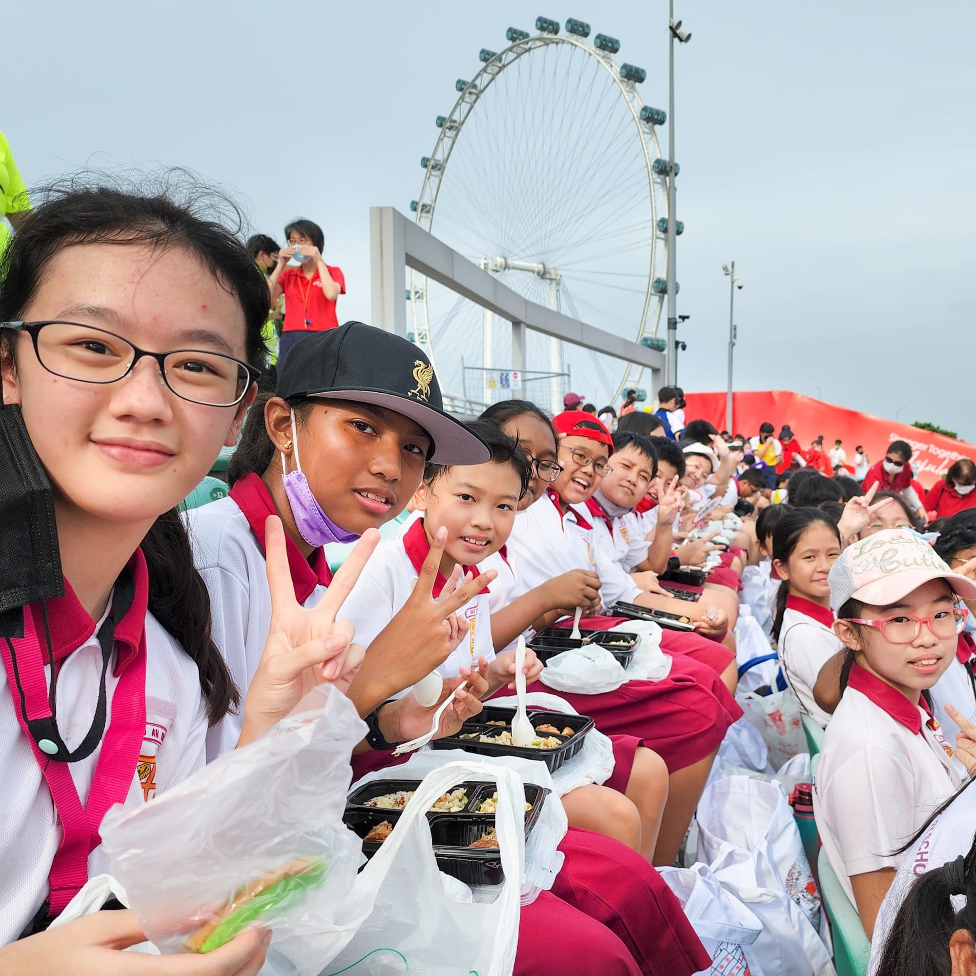 A large group of school children in uniforms eating packed meals outdoors near a large ferris wheel.