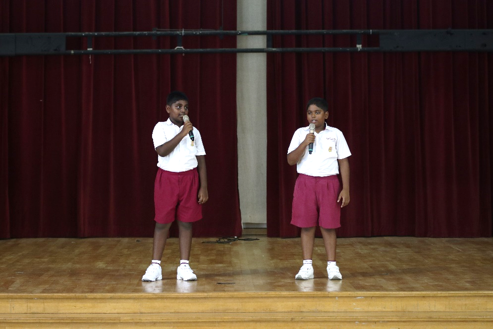 Two children in white shirts and red shorts hold microphones on a wooden stage with a red curtain backdrop.