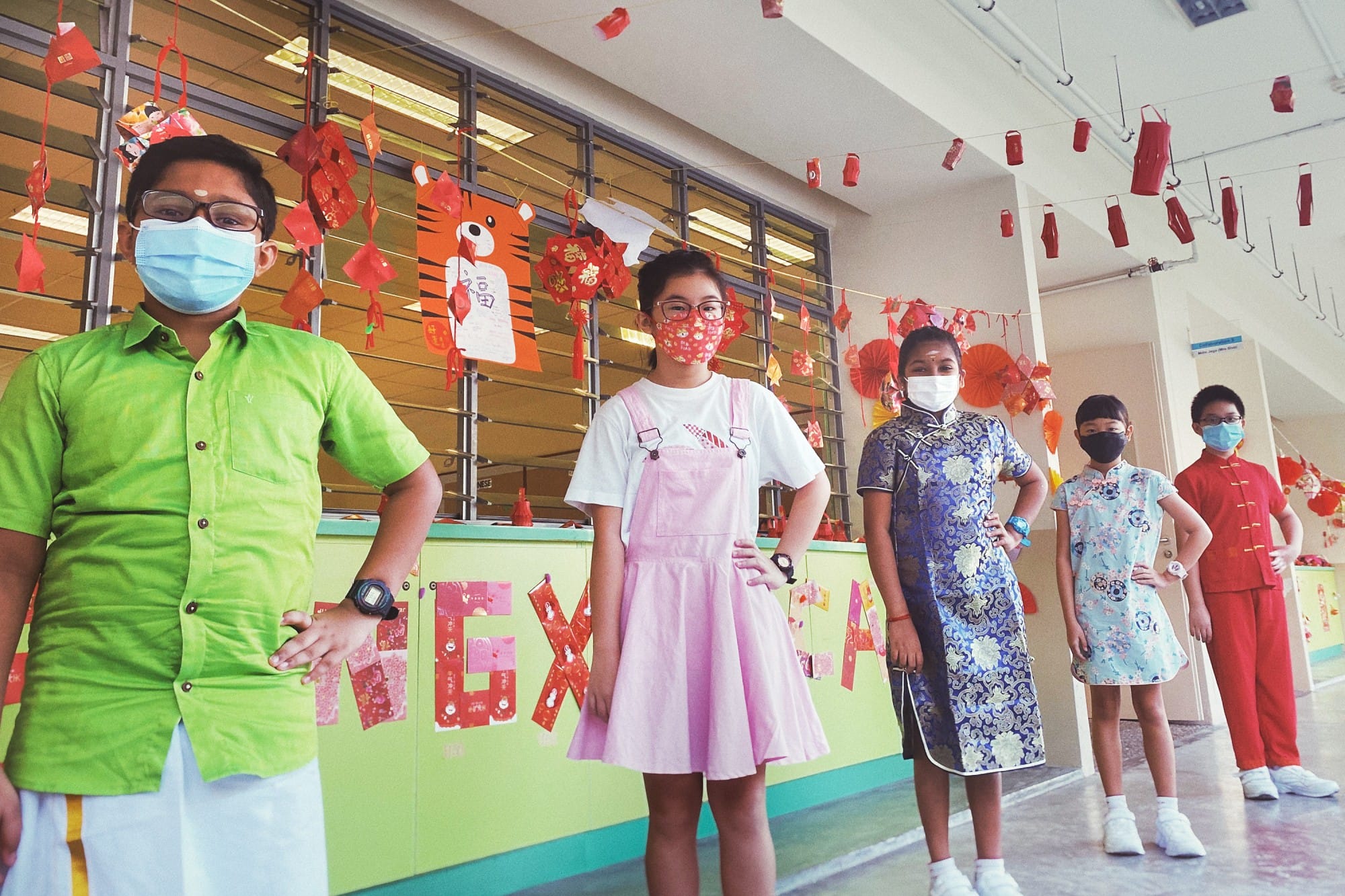 Five children wearing face masks stand in a school hallway decorated with red Chinese New Year lanterns and paper crafts.