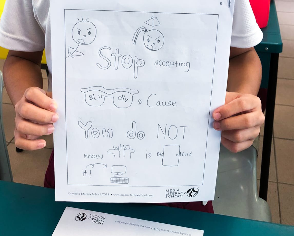 Child holds a hand-drawn worksheet reading "Stop accepting blindly cause you do not know who is behind it."