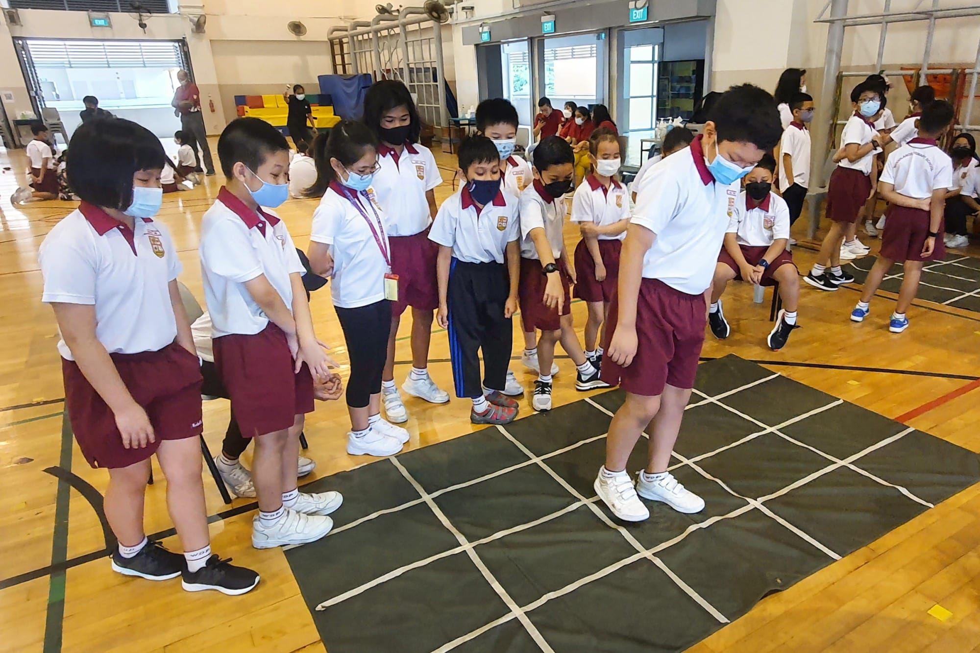 Students in white and maroon uniforms wearing face masks stand around a grid mat on a gymnasium floor.