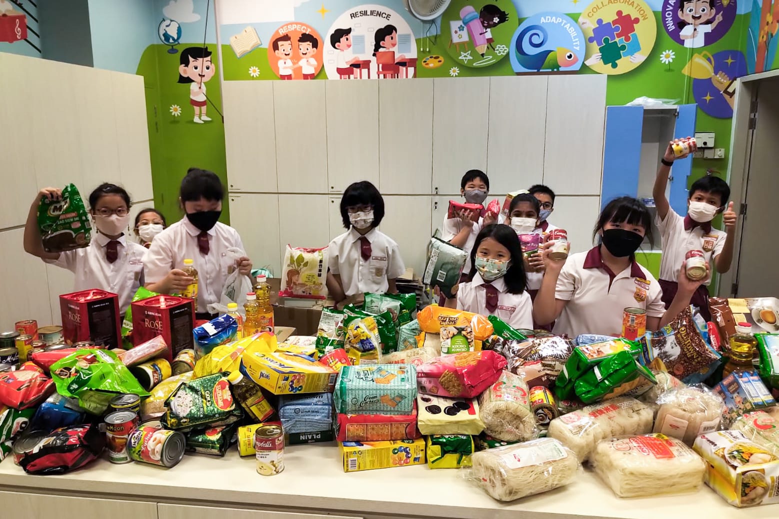 Students in school uniforms and masks stand behind a table piled with donated food items in a colorful classroom.