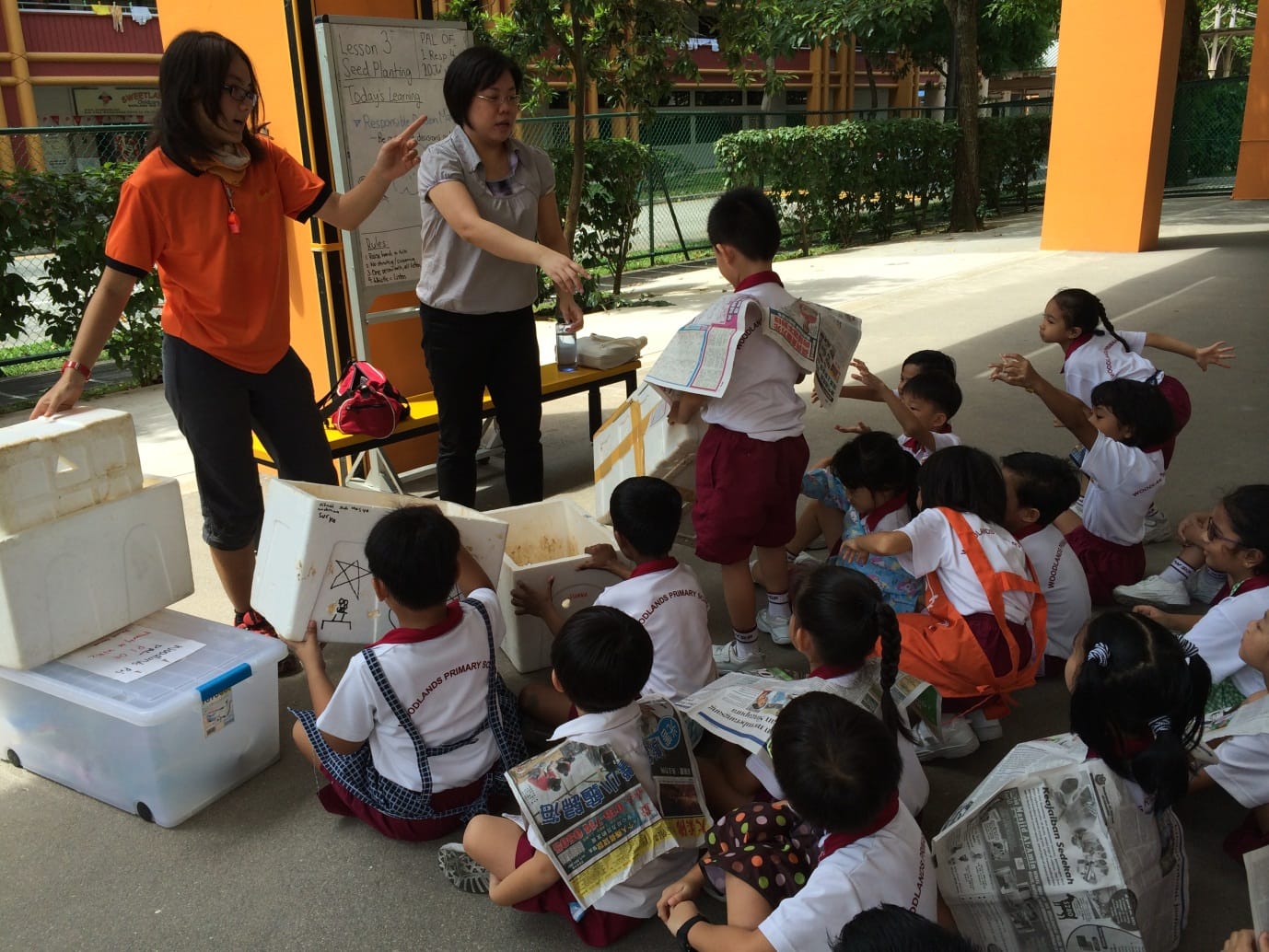 Two teachers instruct young students in school uniforms outdoors during a seed planting lesson with boxes and newspapers.