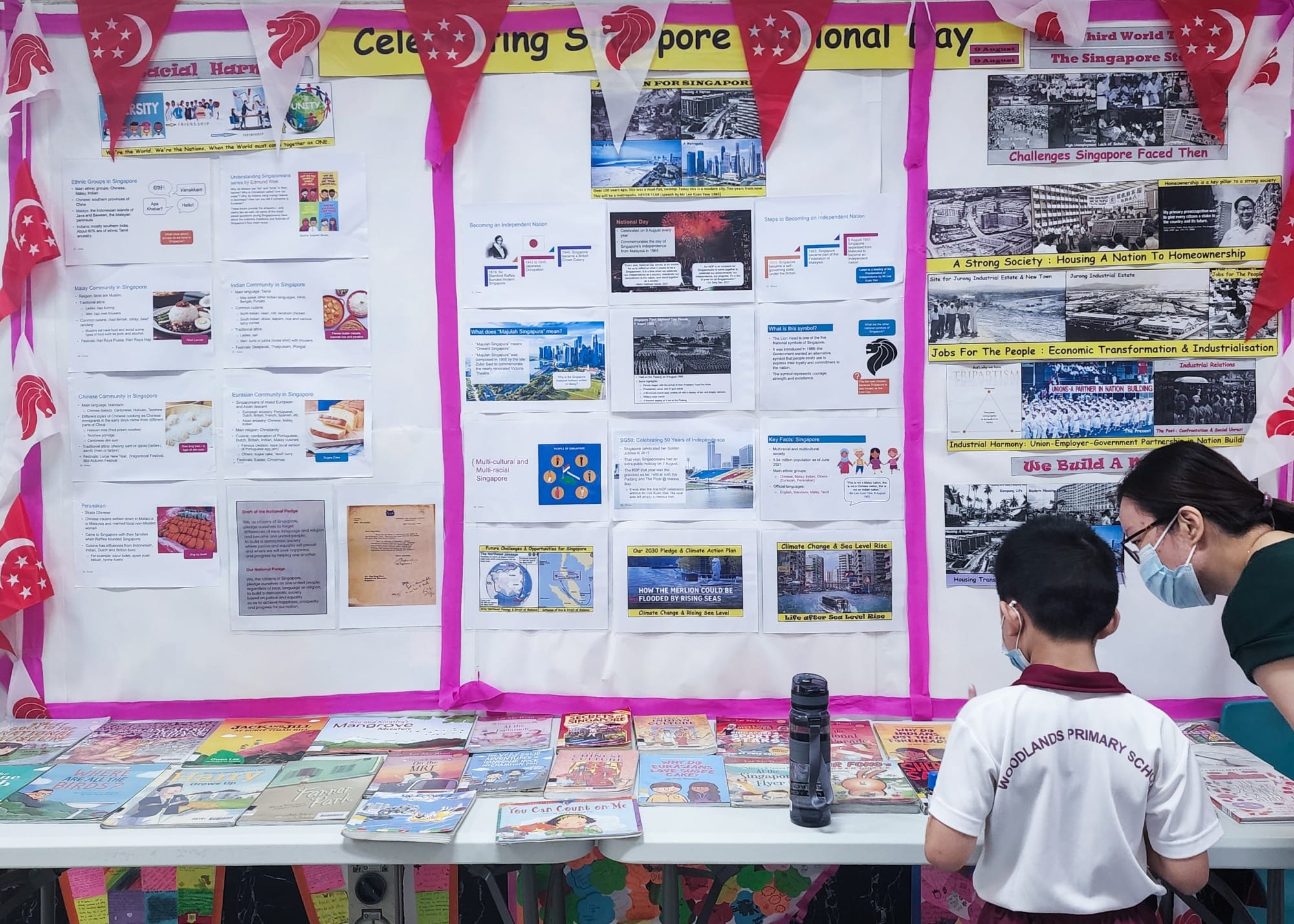 A Woodlands Primary School student and adult view a Singapore National Day display board with books on a table.