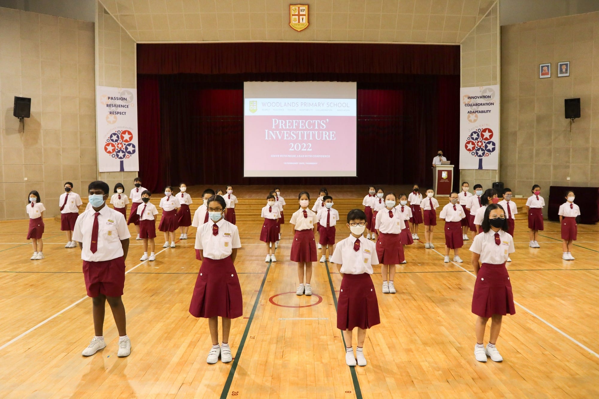 Students in white and red uniforms wearing masks stand in a school hall for Woodlands Primary School Prefects Investiture 2022.