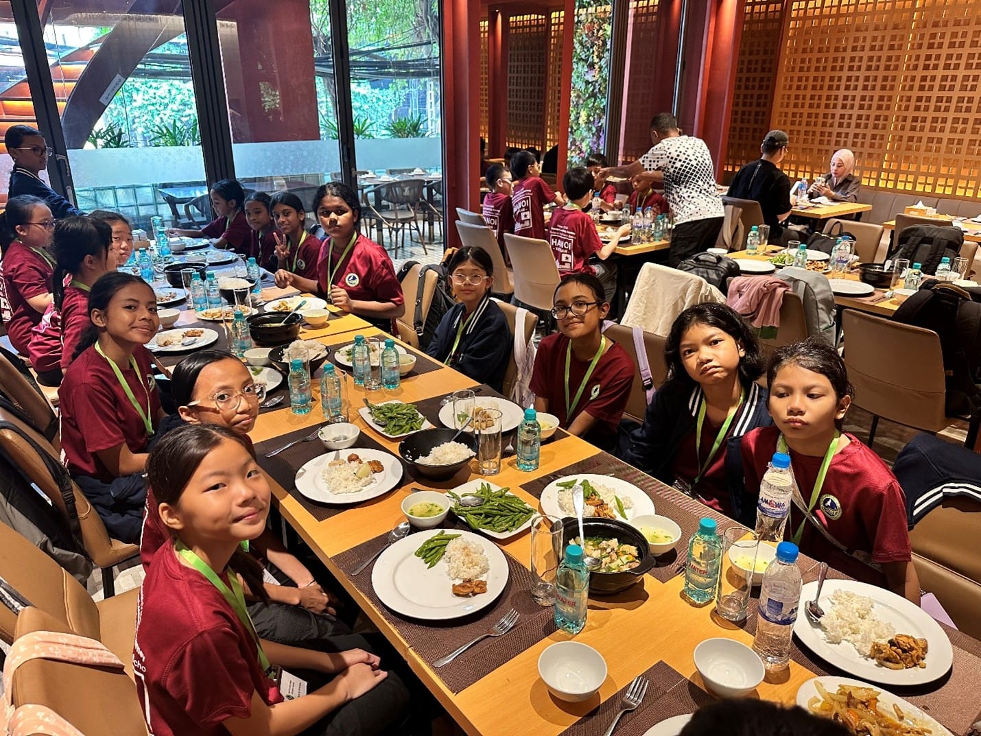 Students from Woodlands Ring Primary School in maroon programme T-shirts enjoy a group meal at a restaurant in Hanoi, with dishes of rice, vegetables, stir-fried meat, and soup served family-style across long dining tables.