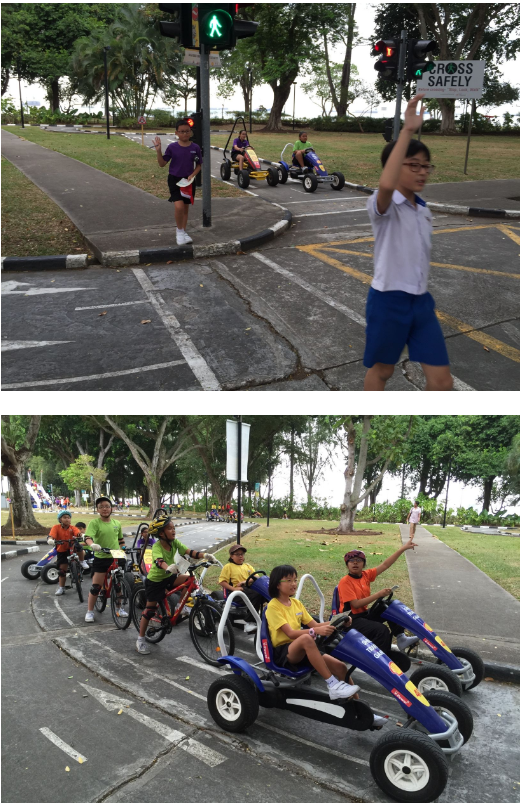 Children practice road safety near a crosswalk with traffic lights, and ride pedal karts and bicycles on a path.
