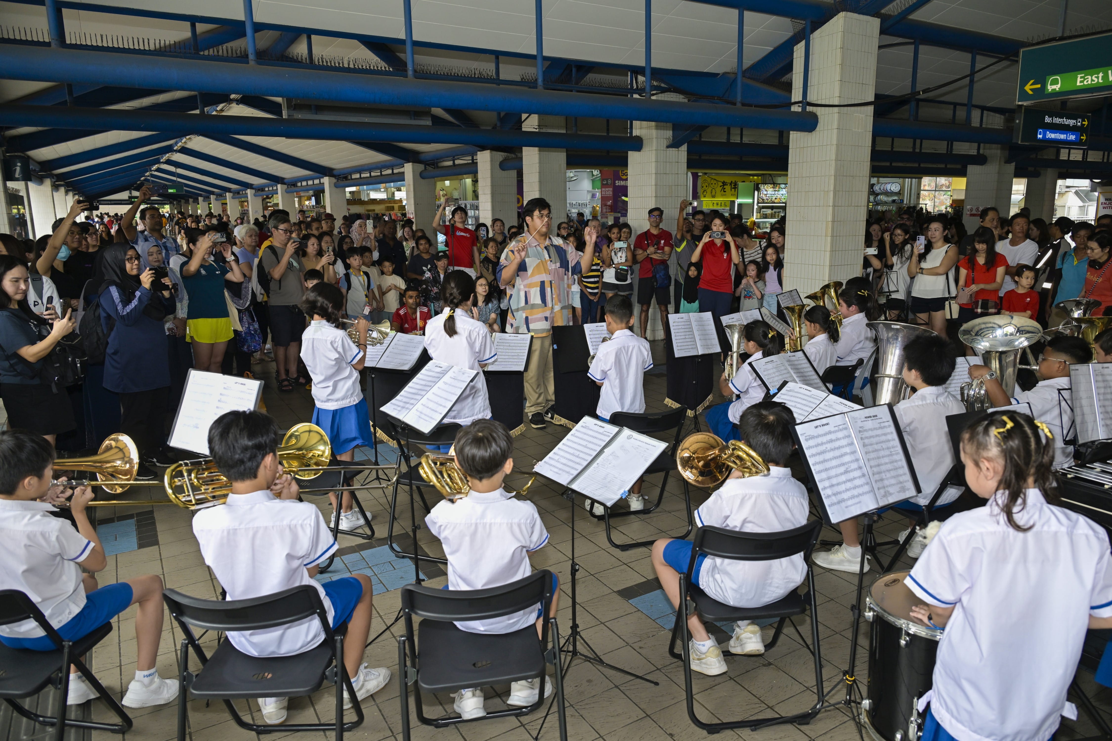 Children in white uniforms play brass instruments outdoors at a transit hub, led by a conductor before a large crowd.