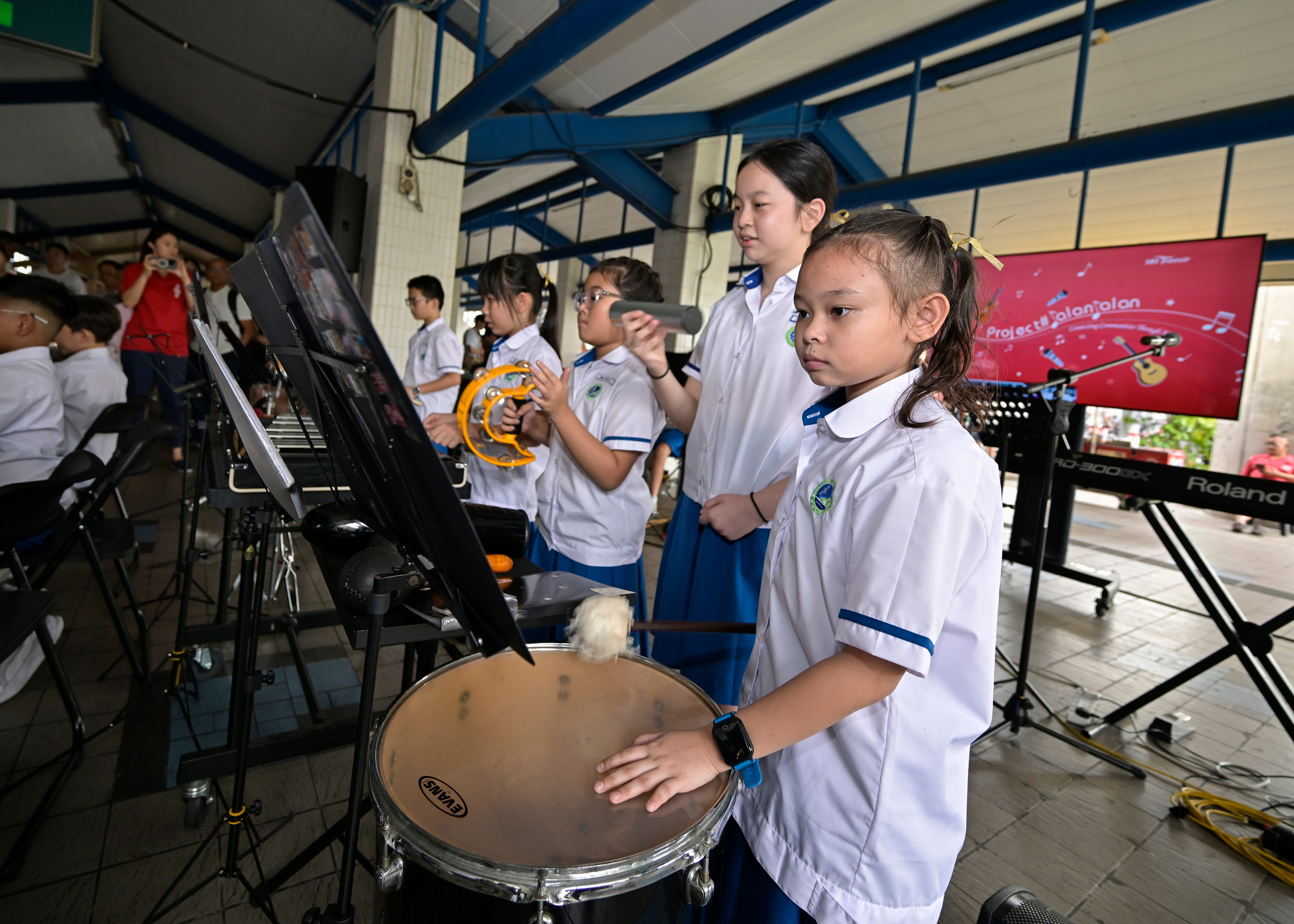 Schoolchildren in white uniforms play percussion, one striking a drum as others hold a mic and tambourine.