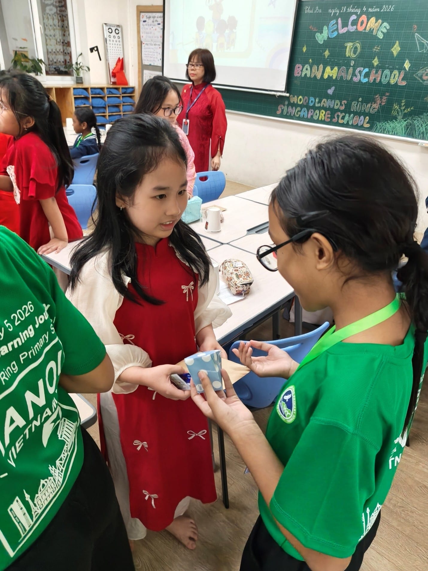 Woodlands Ring Primary School student in a green uniform exchanges a gift with a Ban Mai School student dressed in a traditional Vietnamese ao dai, inside a classroom. A chalkboard in the background reads 'Welcome to Ban Mai School, Woodlands Ring Singapore School'