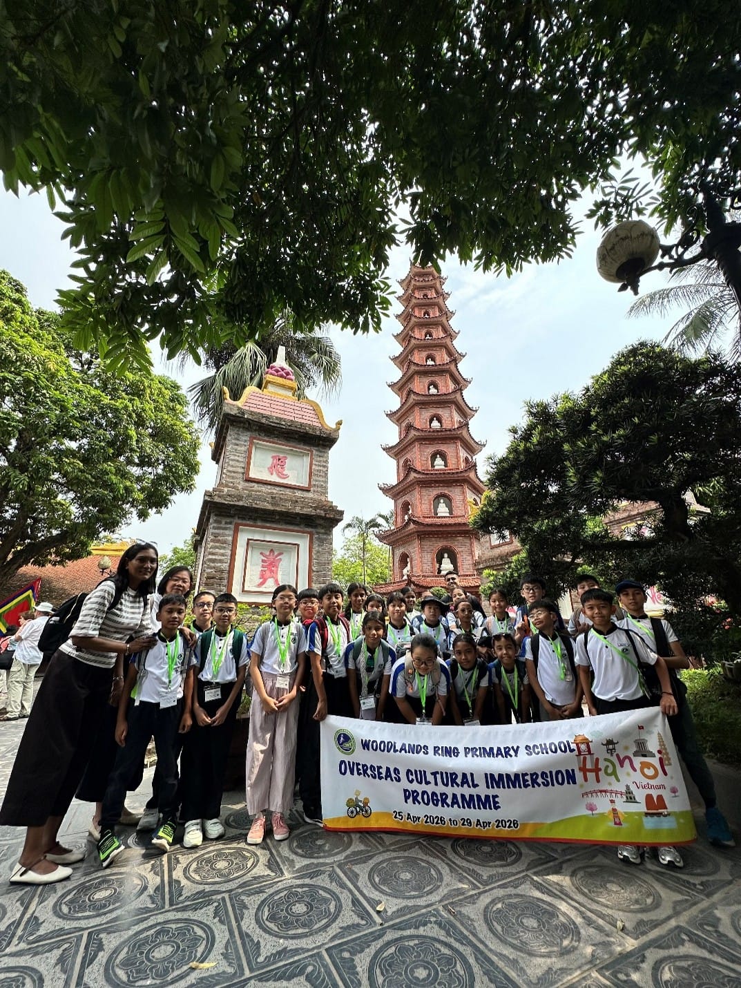 Students,teachers and Principal from Woodlands Ring Primary School pose for a group photo holding the school's Overseas Cultural Immersion Programme banner, with the iconic multi-tiered Tran Quoc Pagoda tower visible in the background.