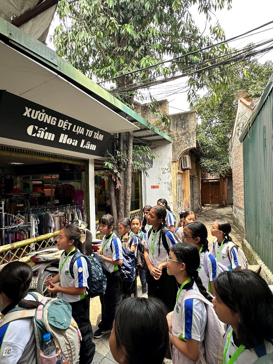Students in school uniforms stand outside the Cam Hoa Lam silk weaving workshop (Xưởng Dệt Lụa Tơ Tằm) in Hanoi, learning about Vietnam's traditional silk weaving craft.