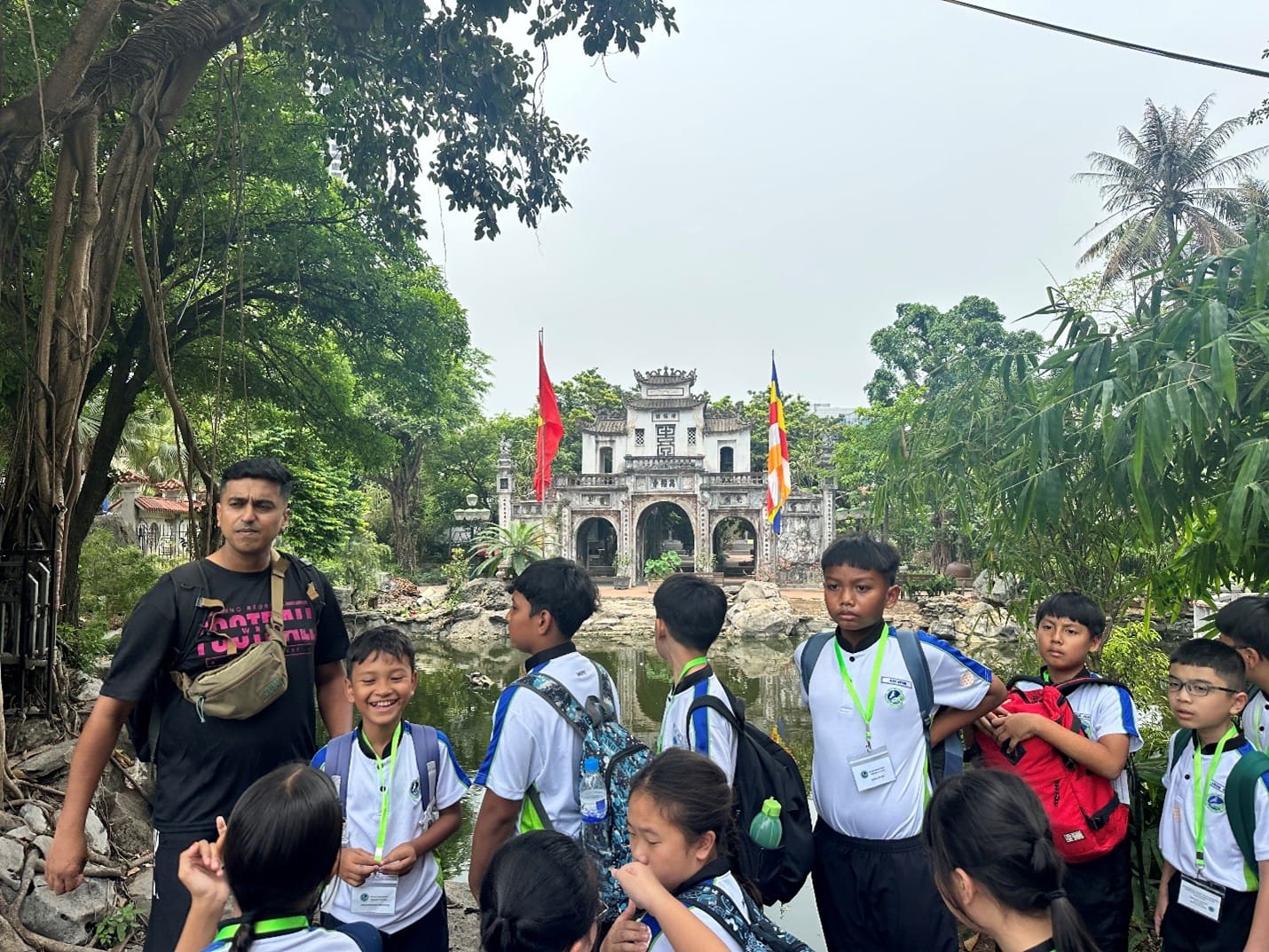 Students and a teacher pause by a tranquil pond with a traditional Vietnamese temple gate reflected in the water, surrounded by lush greenery during their cultural excursion in Hanoi.