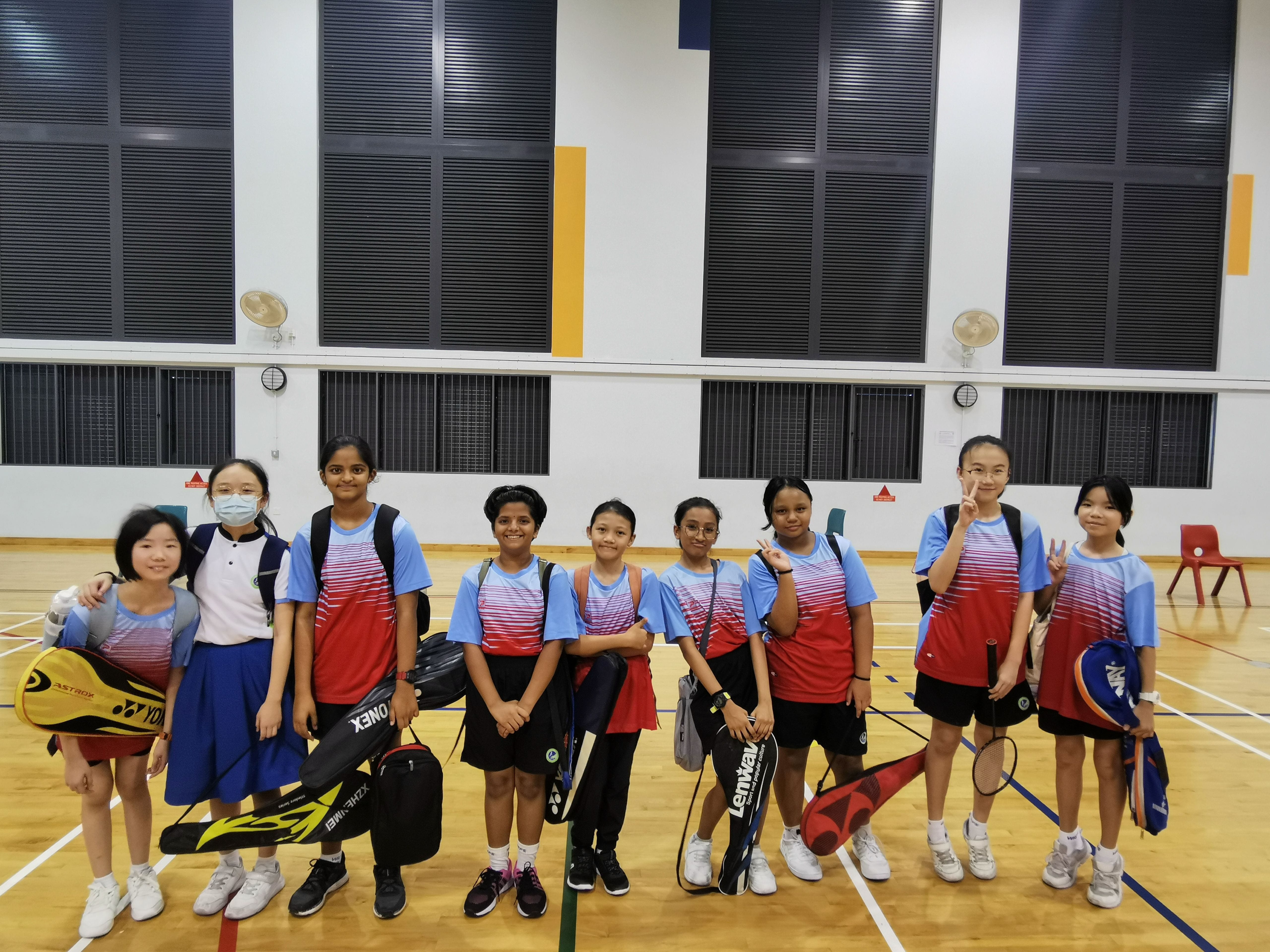 Nine young girls in blue and red sports uniforms pose together on an indoor badminton court, holding rackets and bags.