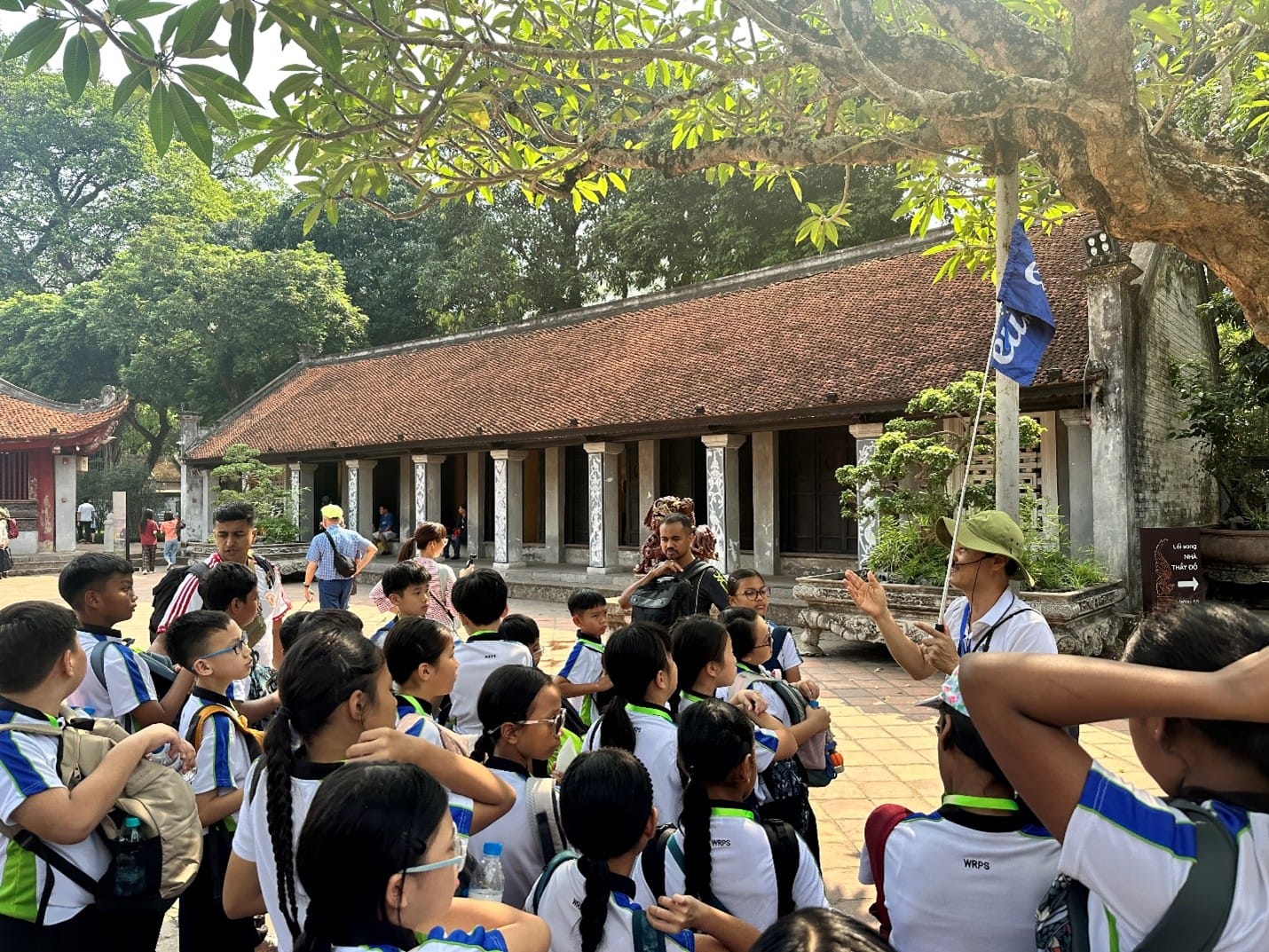 A guide holding a tour flag addresses a large group of students in front of a traditional Vietnamese pavilion with a terracotta-tiled roof at the Temple of Literature in Hanoi.