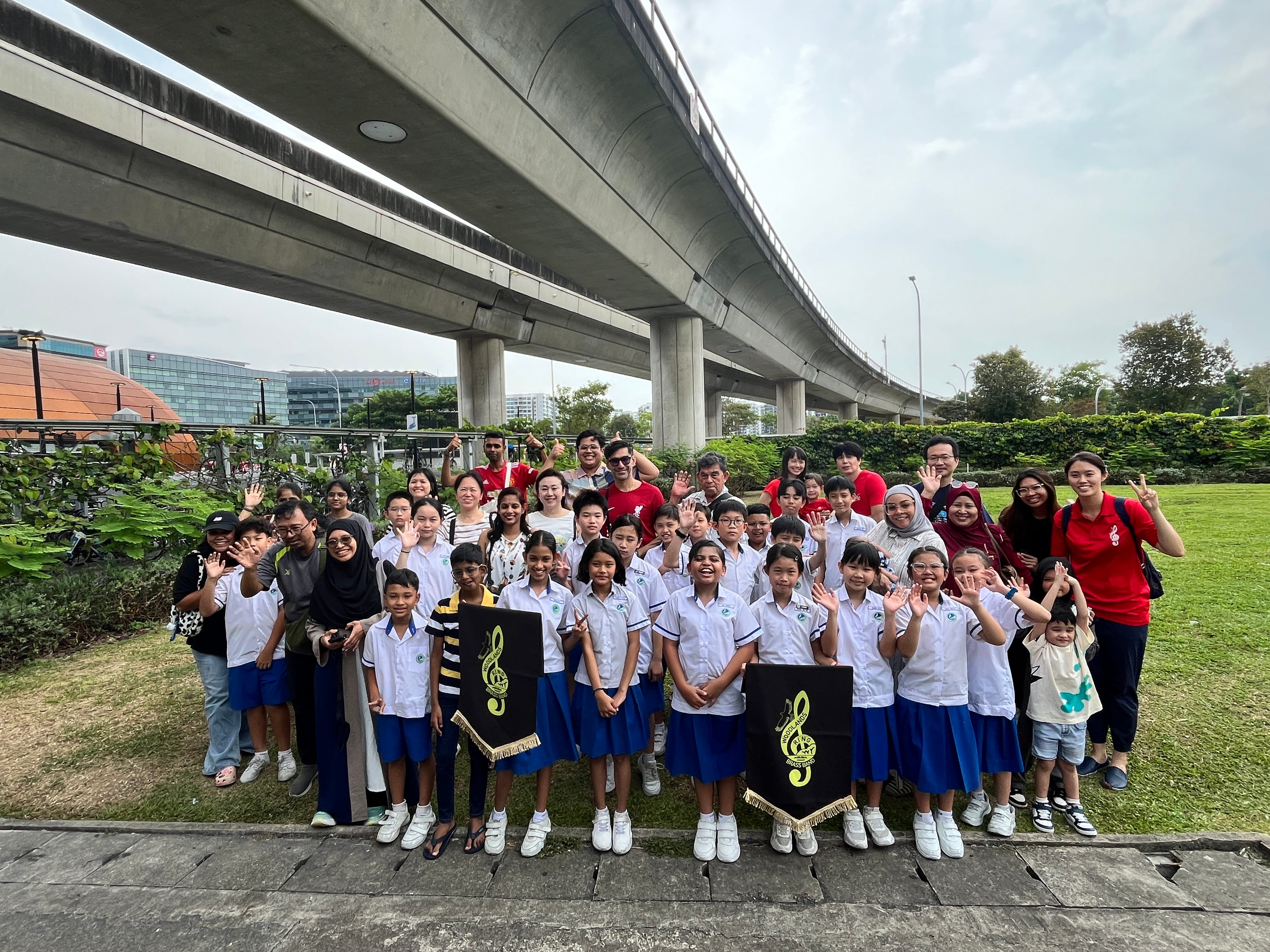 A large group of schoolchildren in white and blue uniforms pose outdoors under an elevated highway, waving and smiling.