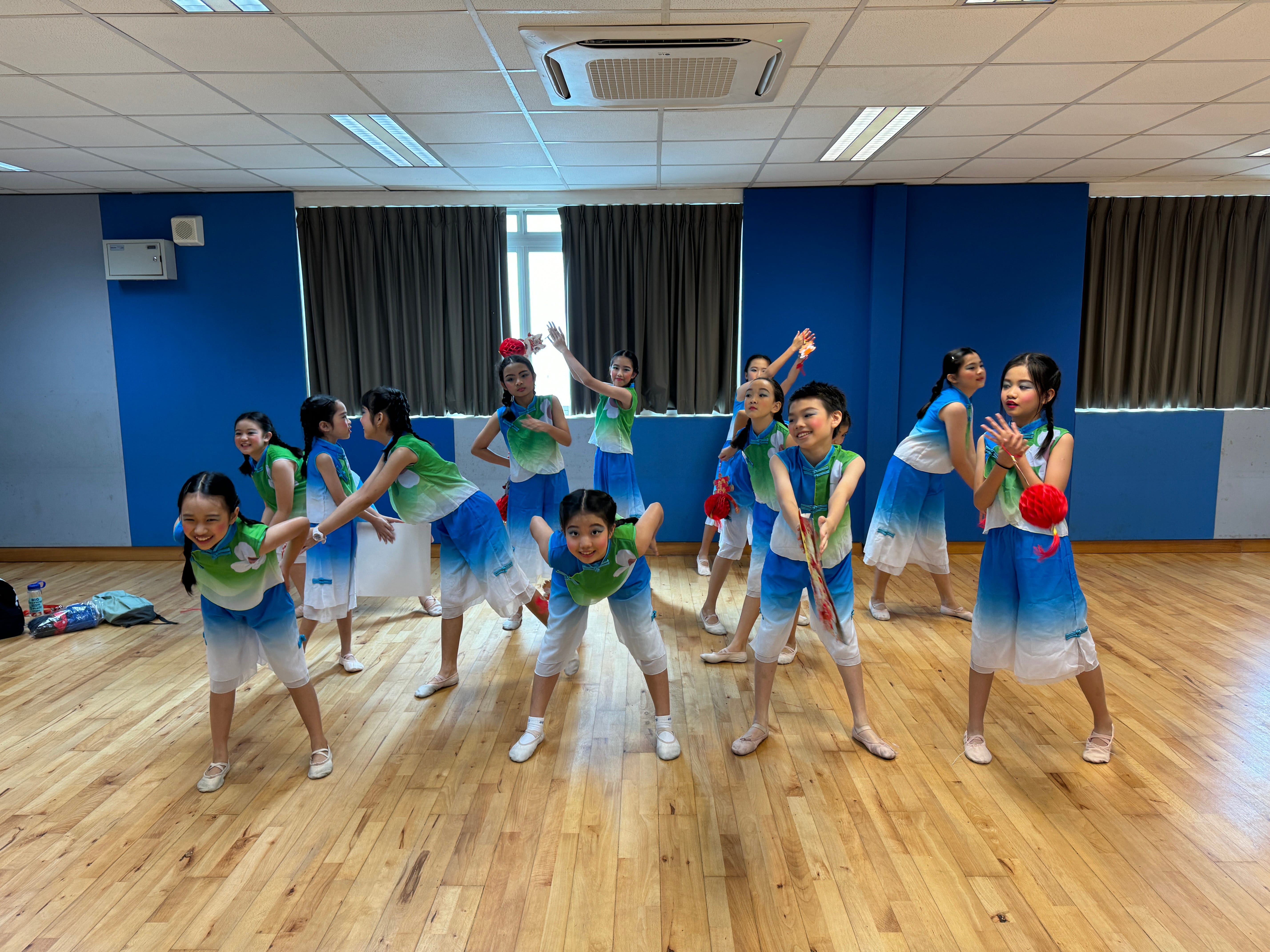 Children in blue-green outfits pose mid-dance in a studio with wooden floor and blue walls.