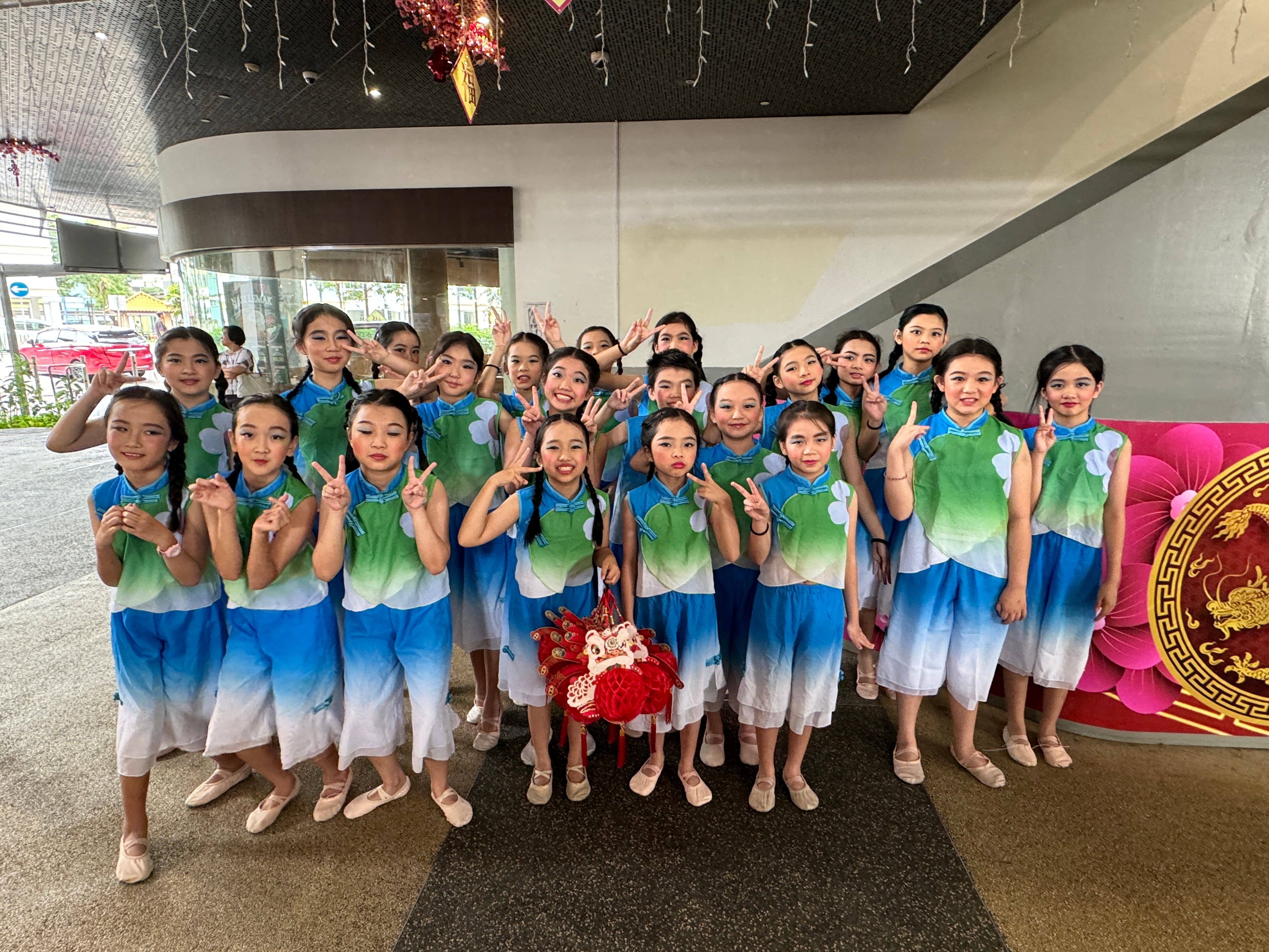 A group of young girls in blue and green Chinese dance costumes pose with peace signs near a festive floral backdrop.