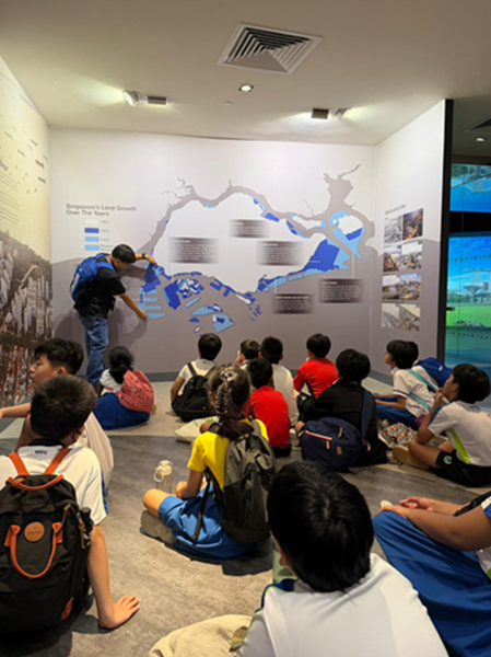 Children sit on the floor in a museum while a guide points to a map of Singapore's land growth on the wall.