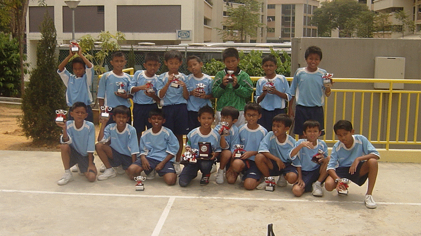 A youth soccer team in light blue uniforms poses in two rows holding trophies outdoors near a yellow railing.