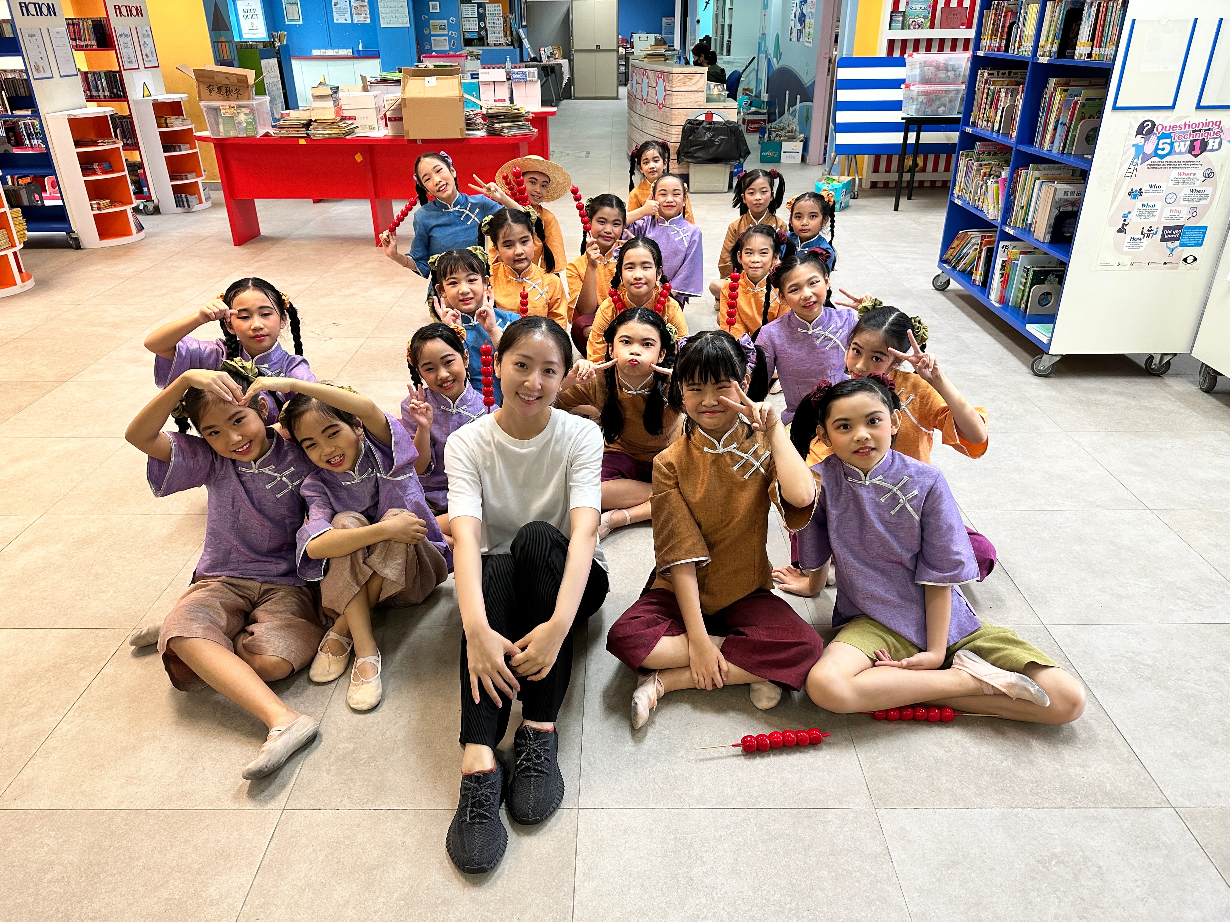 Teacher sits on floor with smiling children in colorful costumes posing in a library among bookshelves.