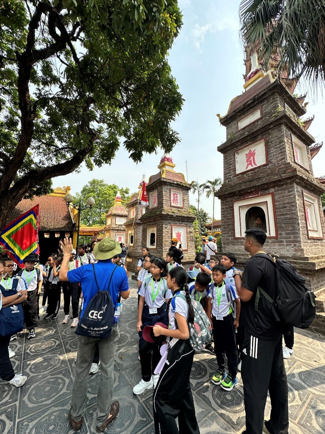 A tour guide in a blue shirt gestures towards ornate stone stupas as students listen attentively during a guided tour at a Vietnamese Buddhist temple complex in Hanoi
