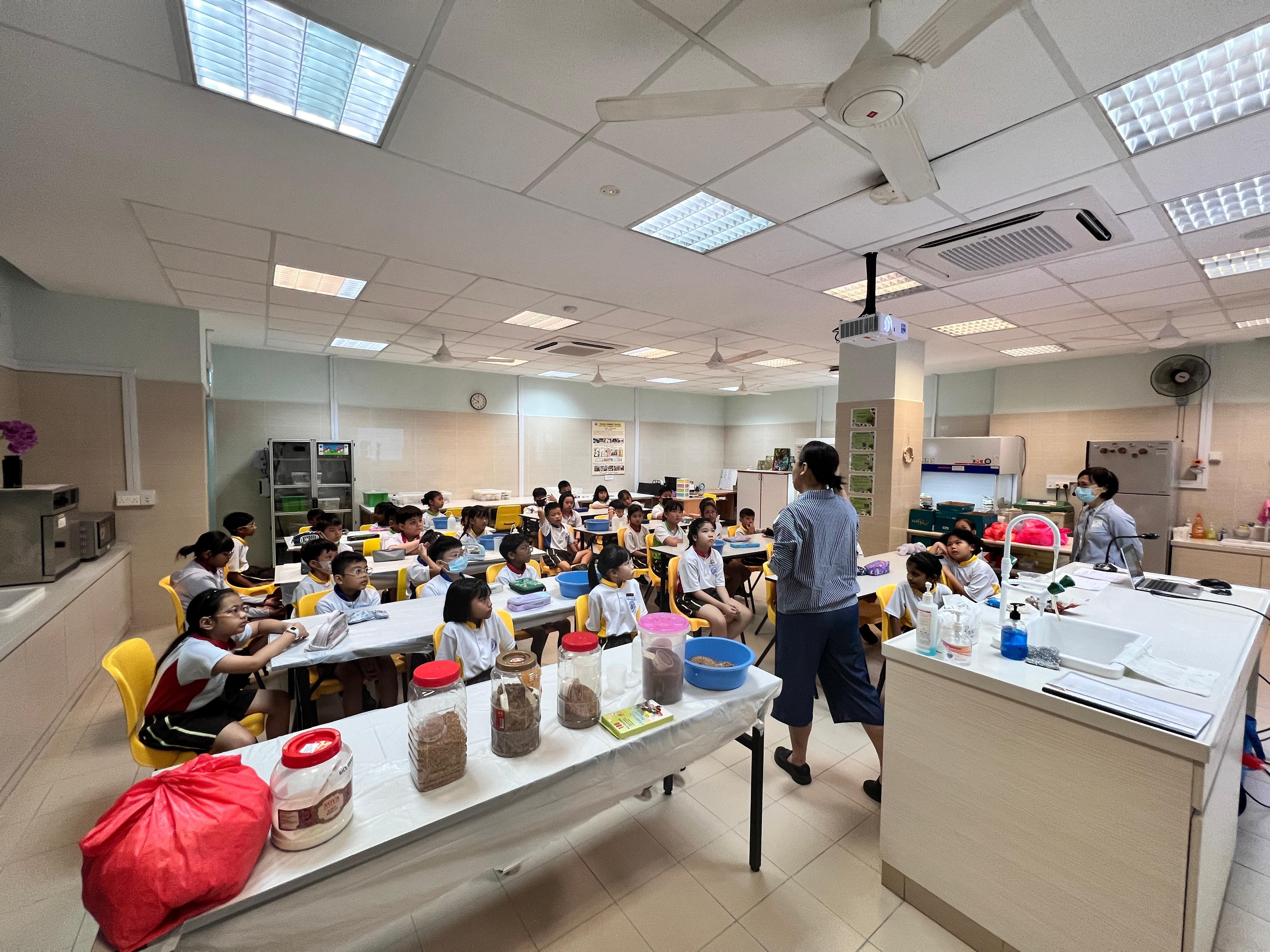 A teacher stands before a large group of young students seated at yellow chairs in a bright classroom with jars on a table.