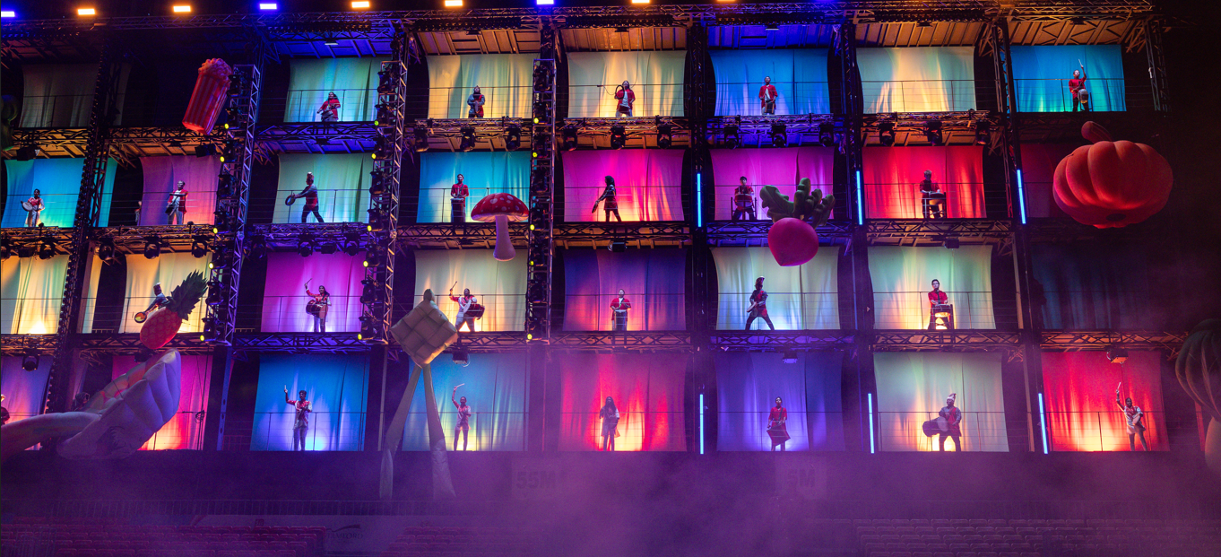 A multi-level stage with performers in each colorful lit compartment, surrounded by giant prop decorations and fog.