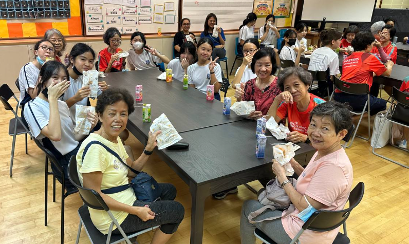 A group of women and girls of various ages sit around a dark table, smiling and holding food packets.