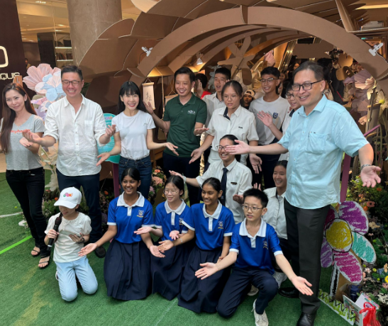 Group of adults and students in blue uniforms posing with open hands under a decorative arch indoors.