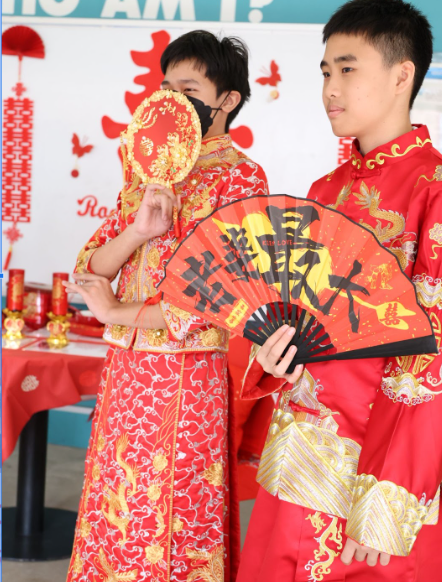 Two young men in red traditional Chinese outfits hold decorative red fans with gold Chinese characters.