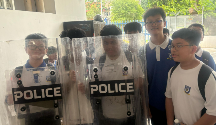 Two students hold large transparent police riot shields while other students stand nearby outdoors.