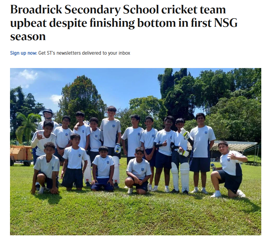 A group of young cricket players in white uniforms posing together on a grass field with trees in the background.
