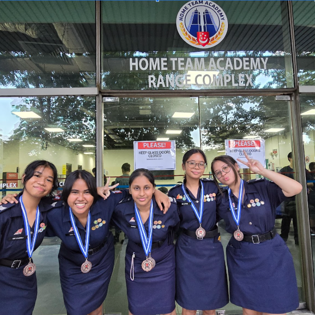 Five girls in navy uniforms wearing medals pose in front of the Home Team Academy Range Complex entrance.