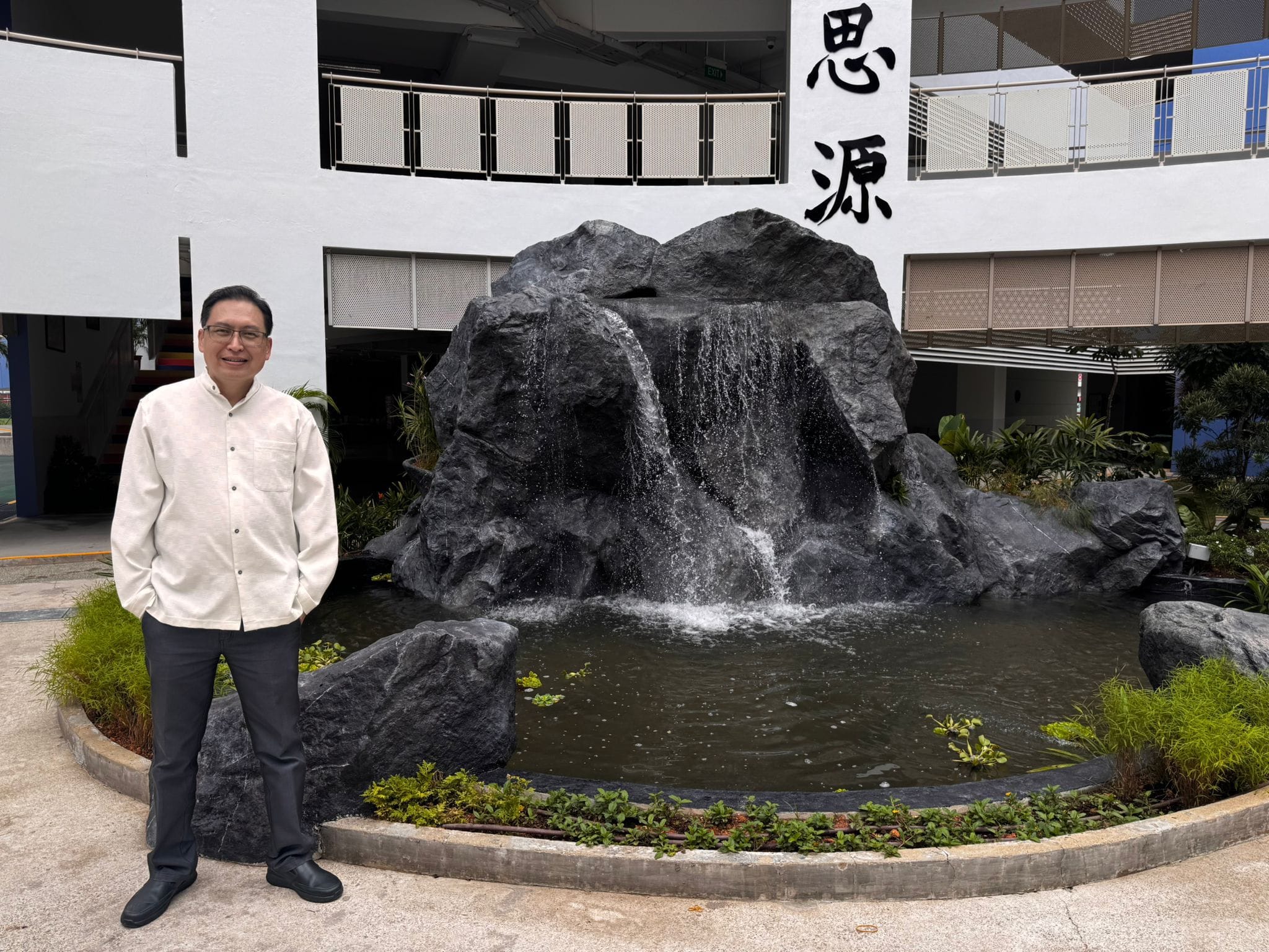 Man in white shirt standing beside a large rock waterfall fountain with Chinese characters on a white building wall.