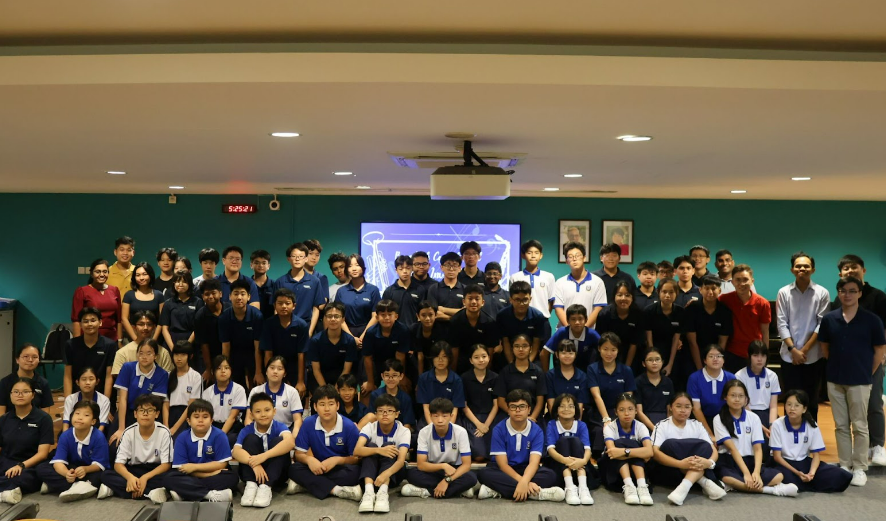 Large group of students in blue and white uniforms posing together indoors in front of a projected screen.