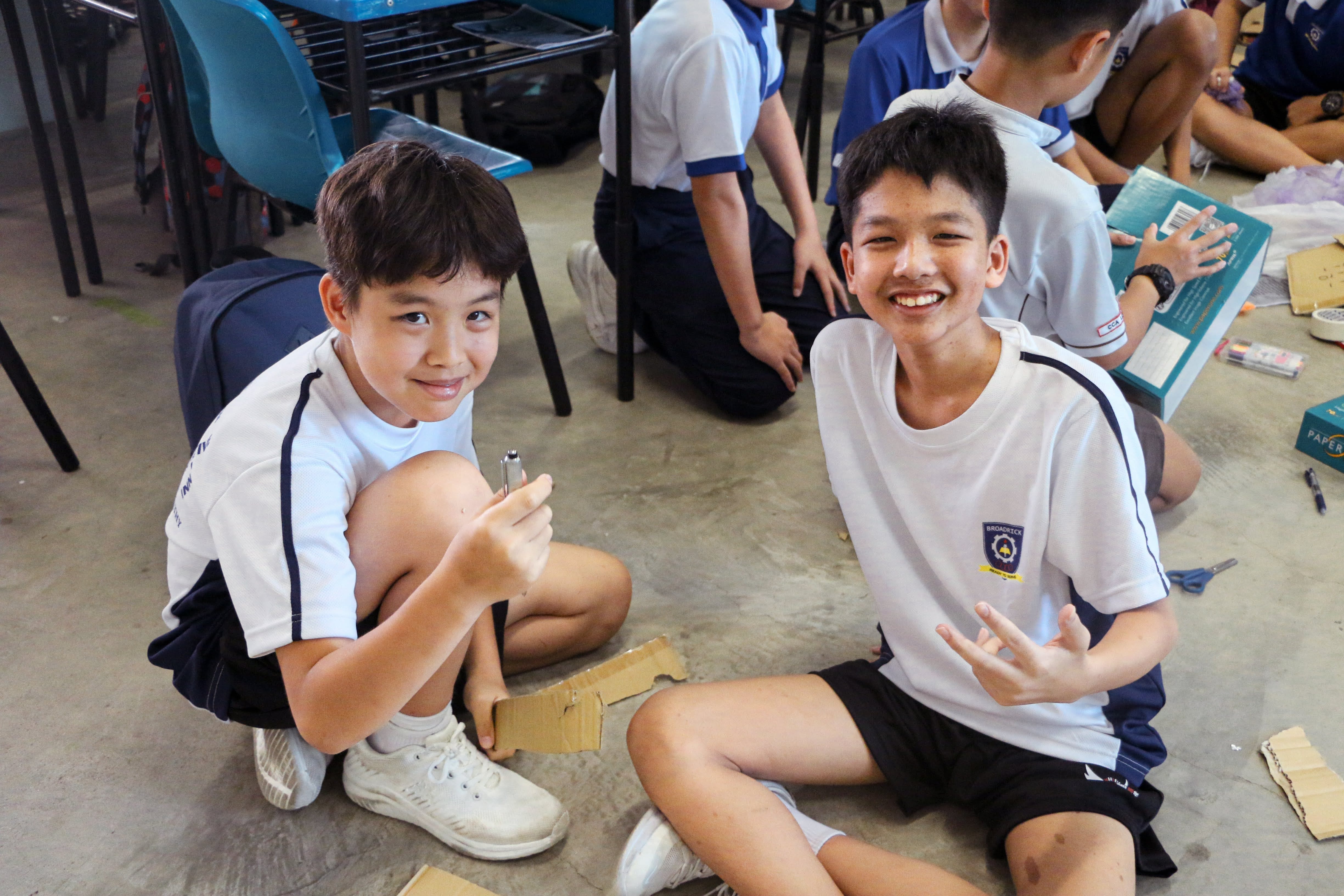 Two smiling schoolboys in white uniforms sit on a classroom floor with cardboard pieces during a group activity.