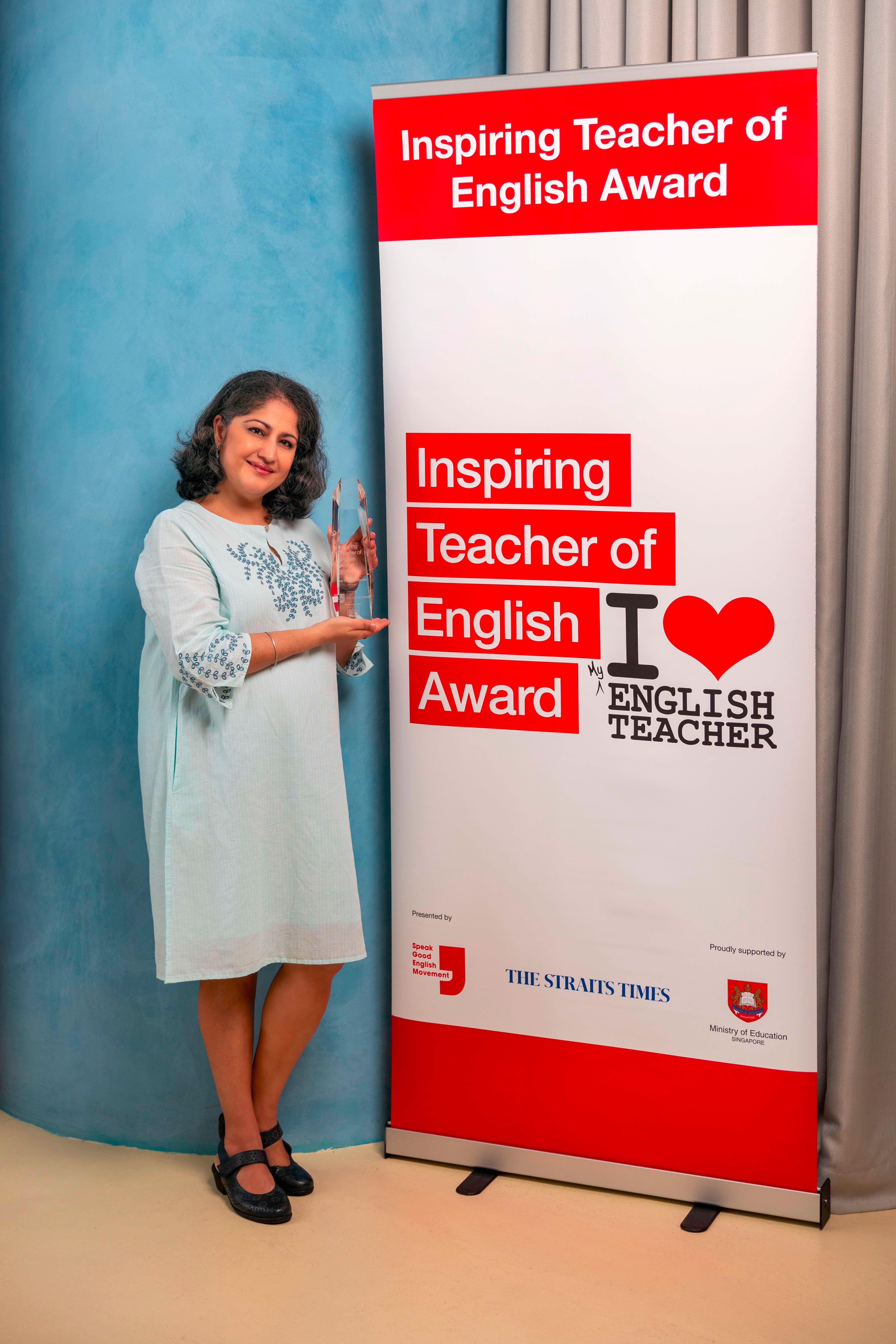 Woman in a light blue dress holds a crystal trophy beside an Inspiring Teacher of English Award banner.