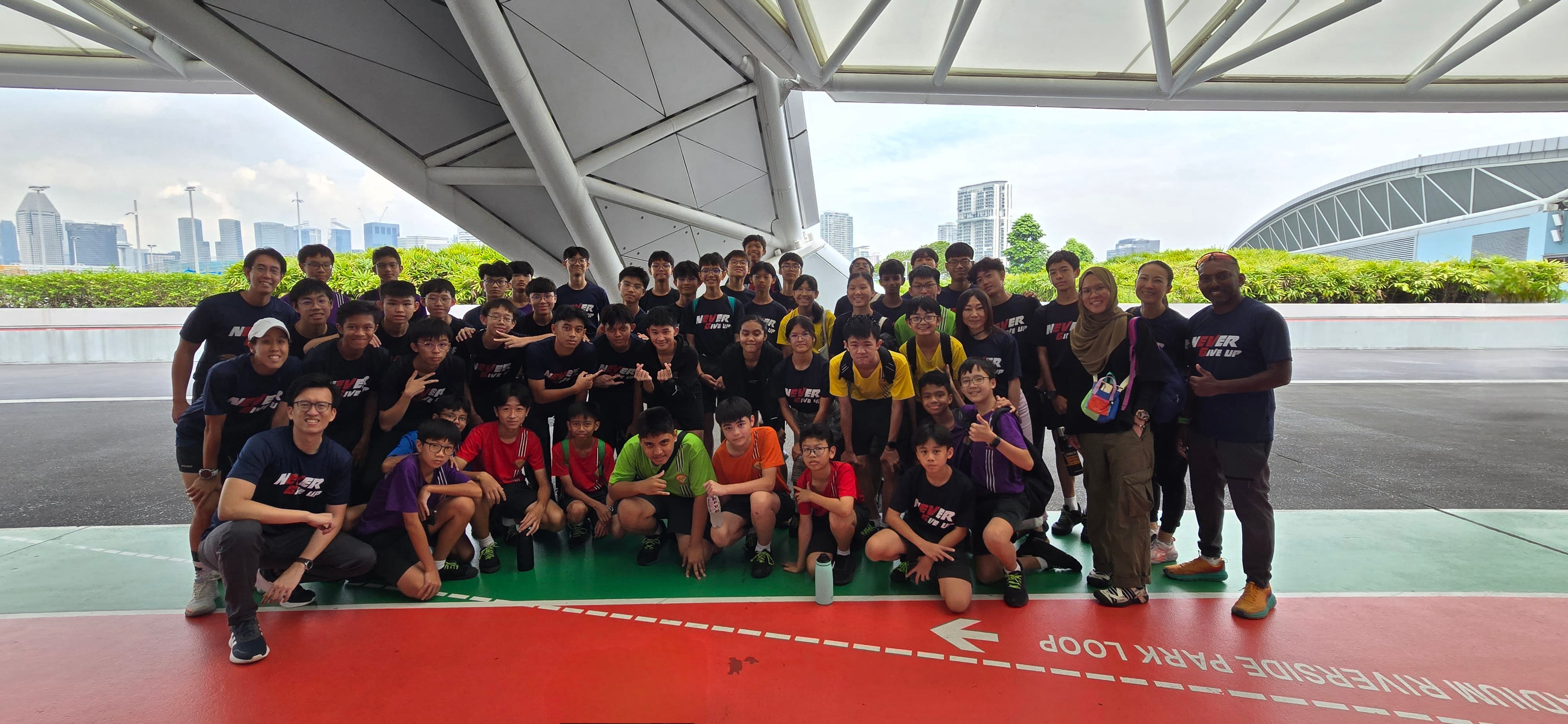 Large group of youth and adults in dark shirts posing outdoors near the Riverside Park Loop path with city skyline behind.