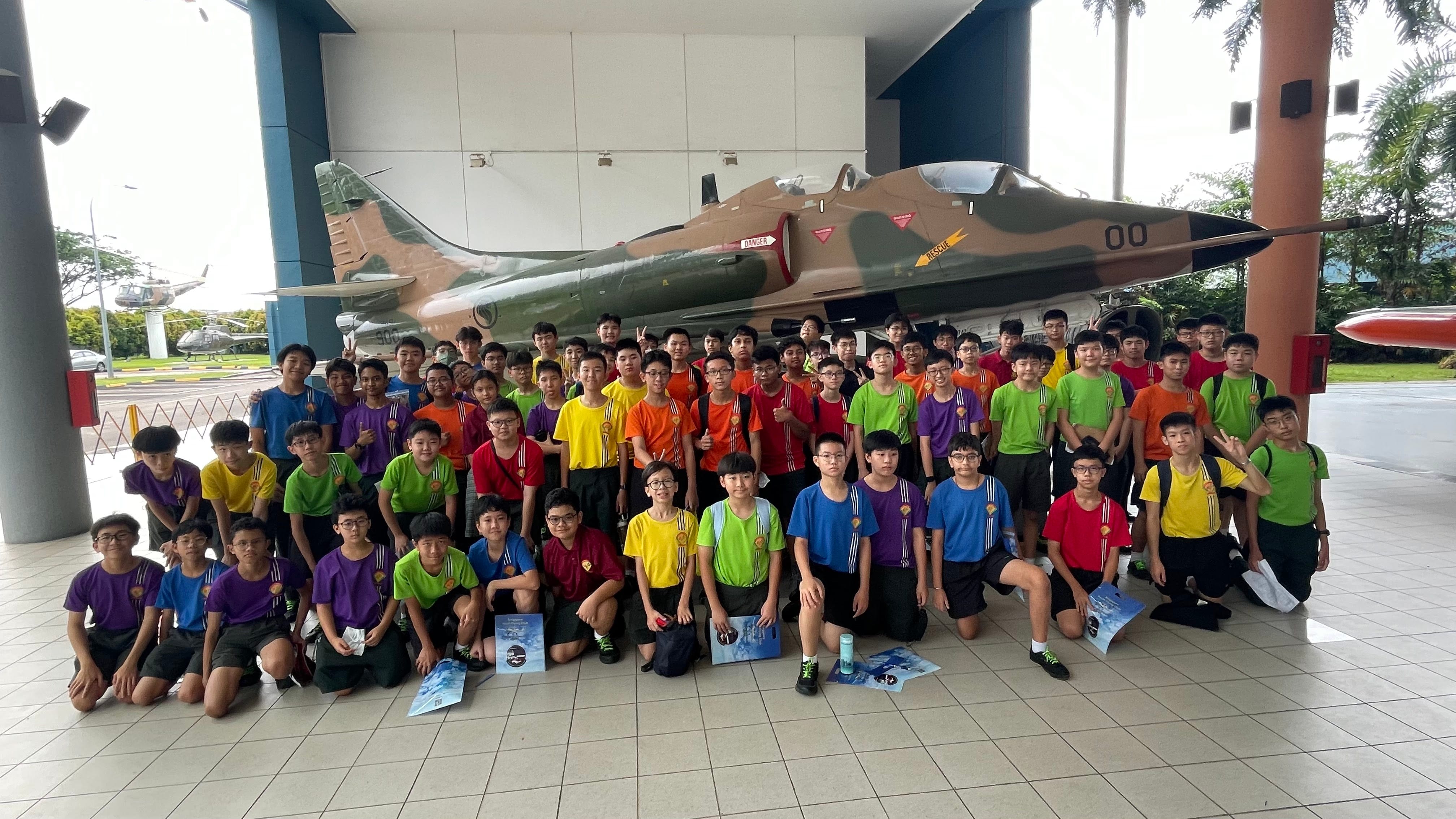A large group of youth in colorful shirts pose together in front of a camouflage military jet on display outdoors.